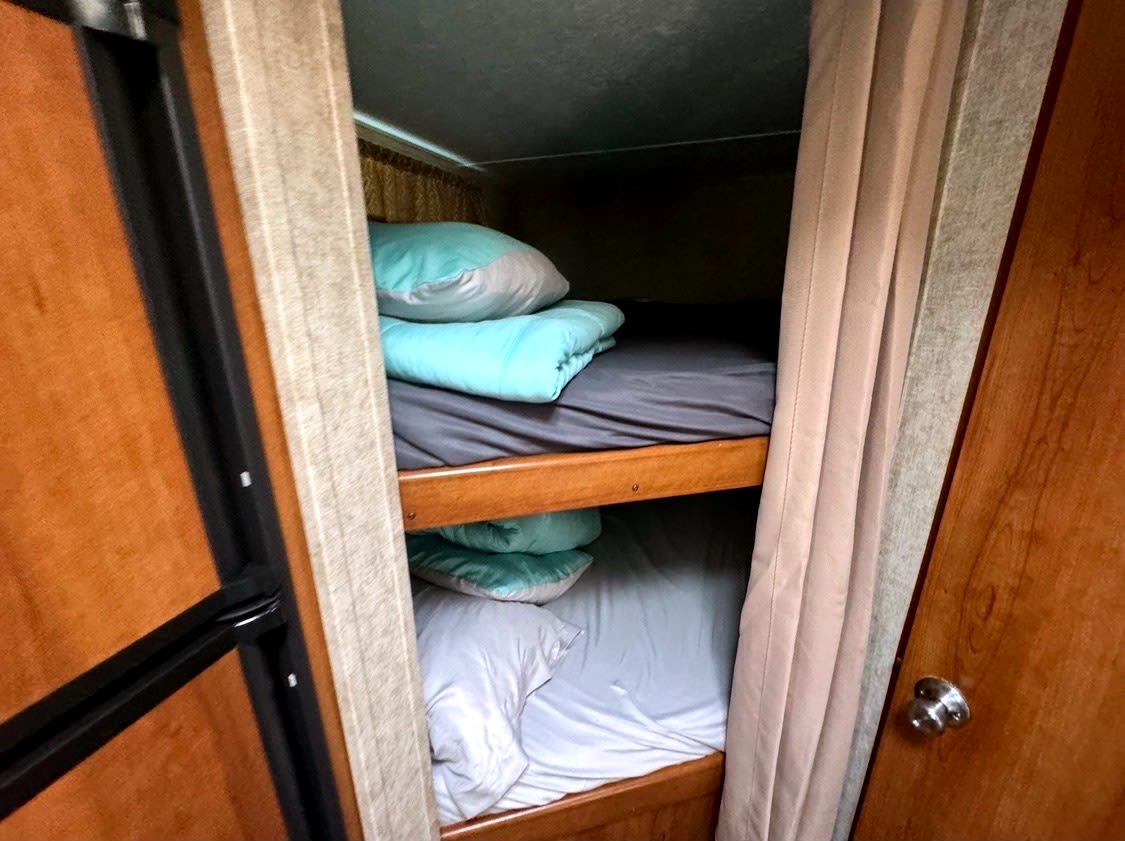 Inside an RV rental: cozy bunk beds with light blue and white bedding, framed by light brown curtains and wood accents.. Forest River Tracer 2016