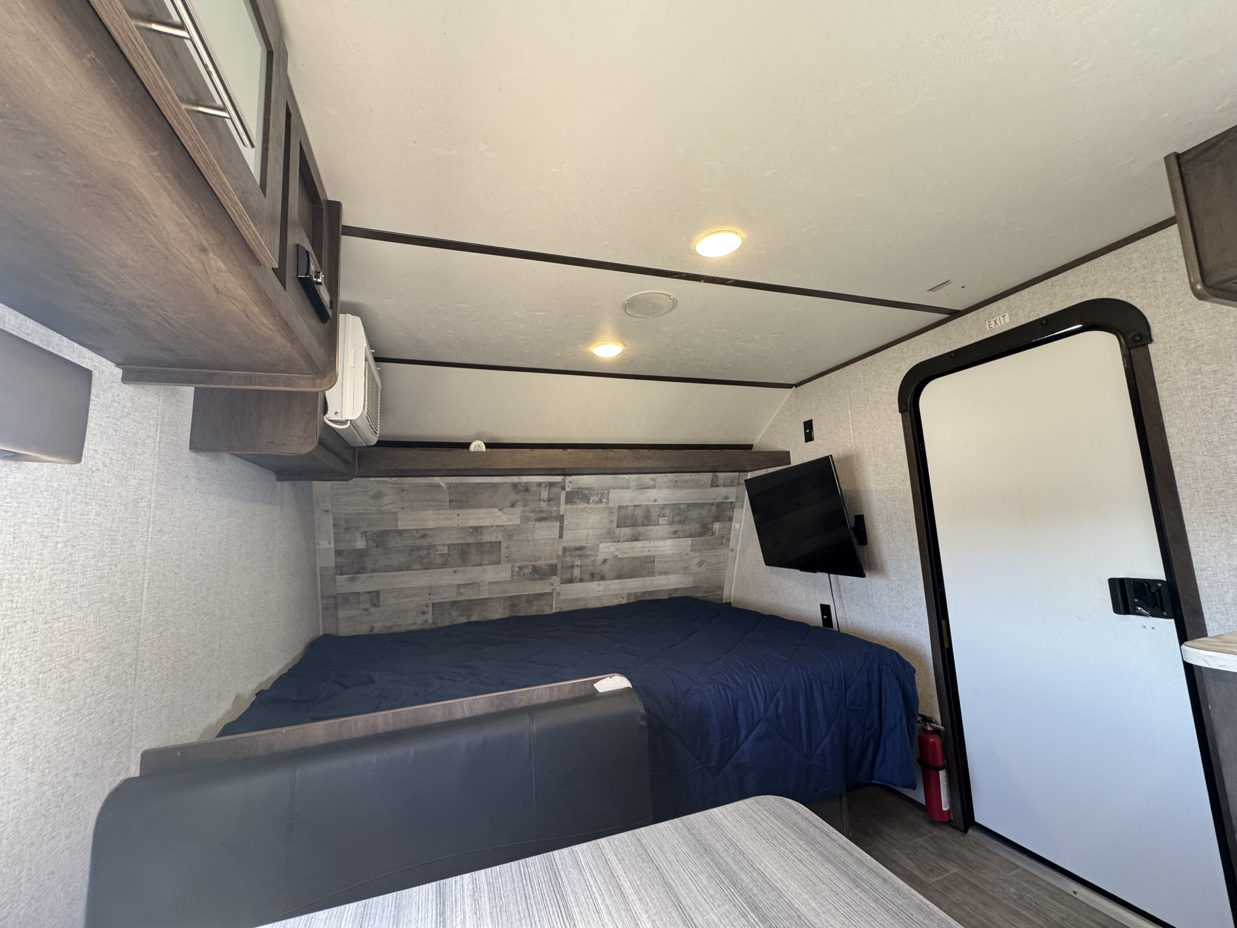 Cozy RV rental interior with a bed, wall-mounted TV, dinette seating, and overhead cabinets. An exit door and fire extinguisher are visible.. Dutchmen Coleman Lantern LT 2021