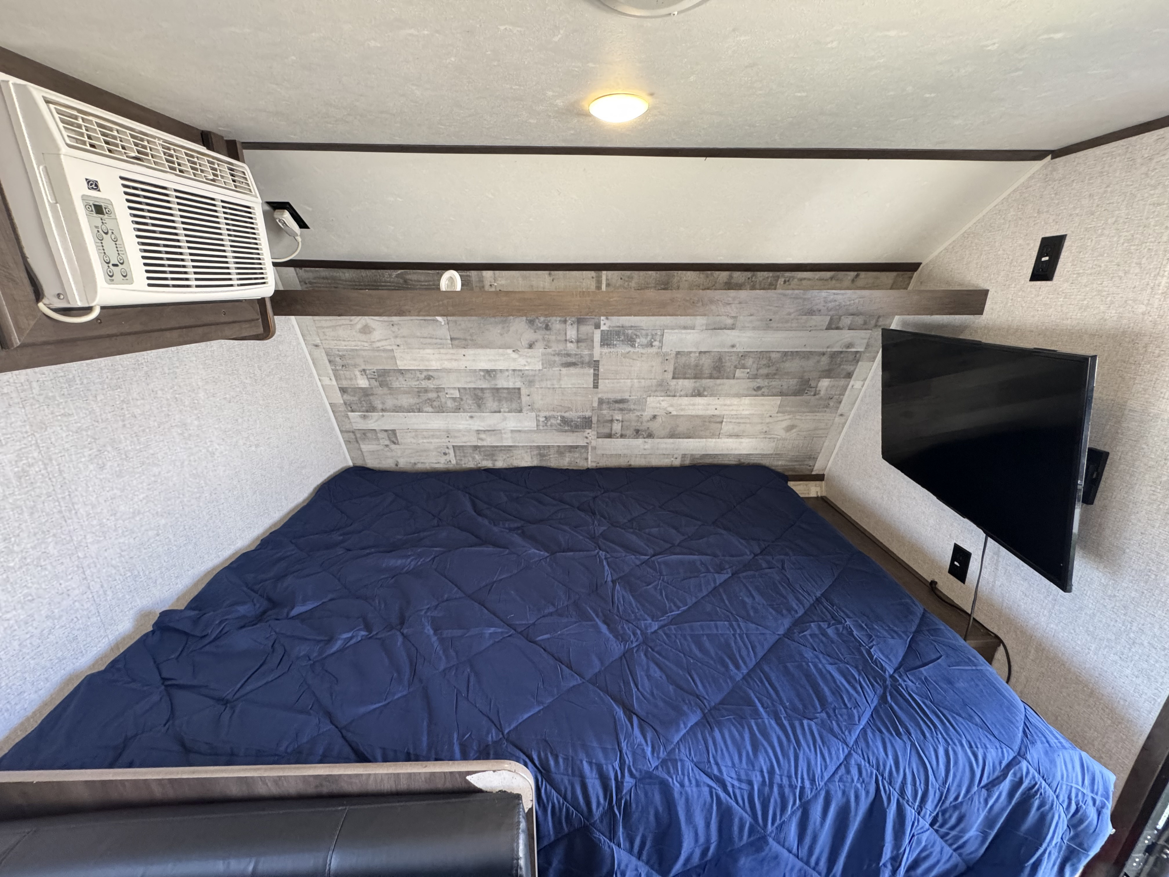Inside an RV rental: a compact bedroom with a navy bed, AC unit, and wall-mounted TV for your comfortable travel accommodation.. Dutchmen Coleman Lantern LT 2021