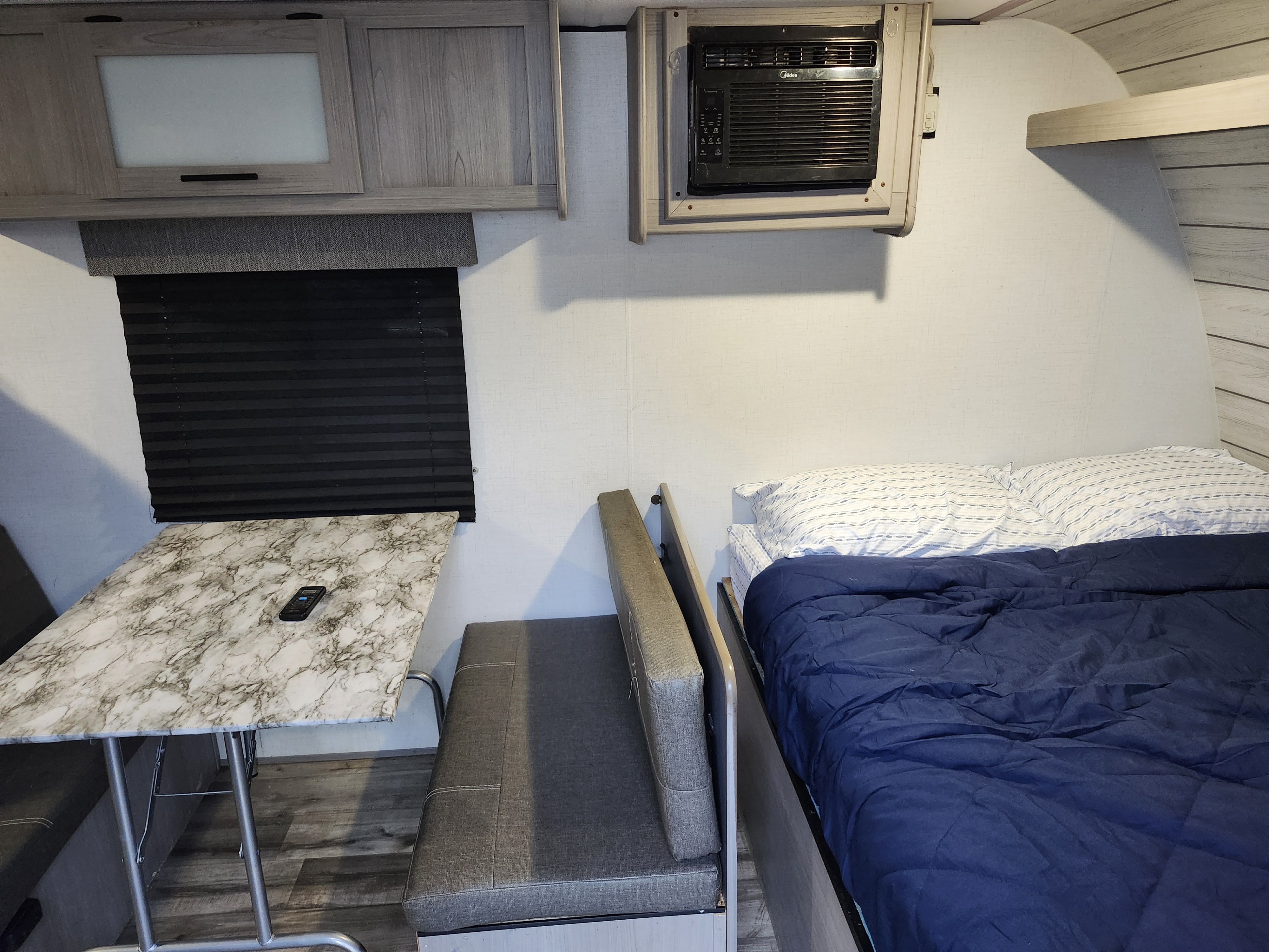 This RV rental interior features a compact dinette with a marble-top table, overhead storage, a cozy bed with blue bedding, and a wall-mounted AC unit.. Dutchmen Coleman 2024