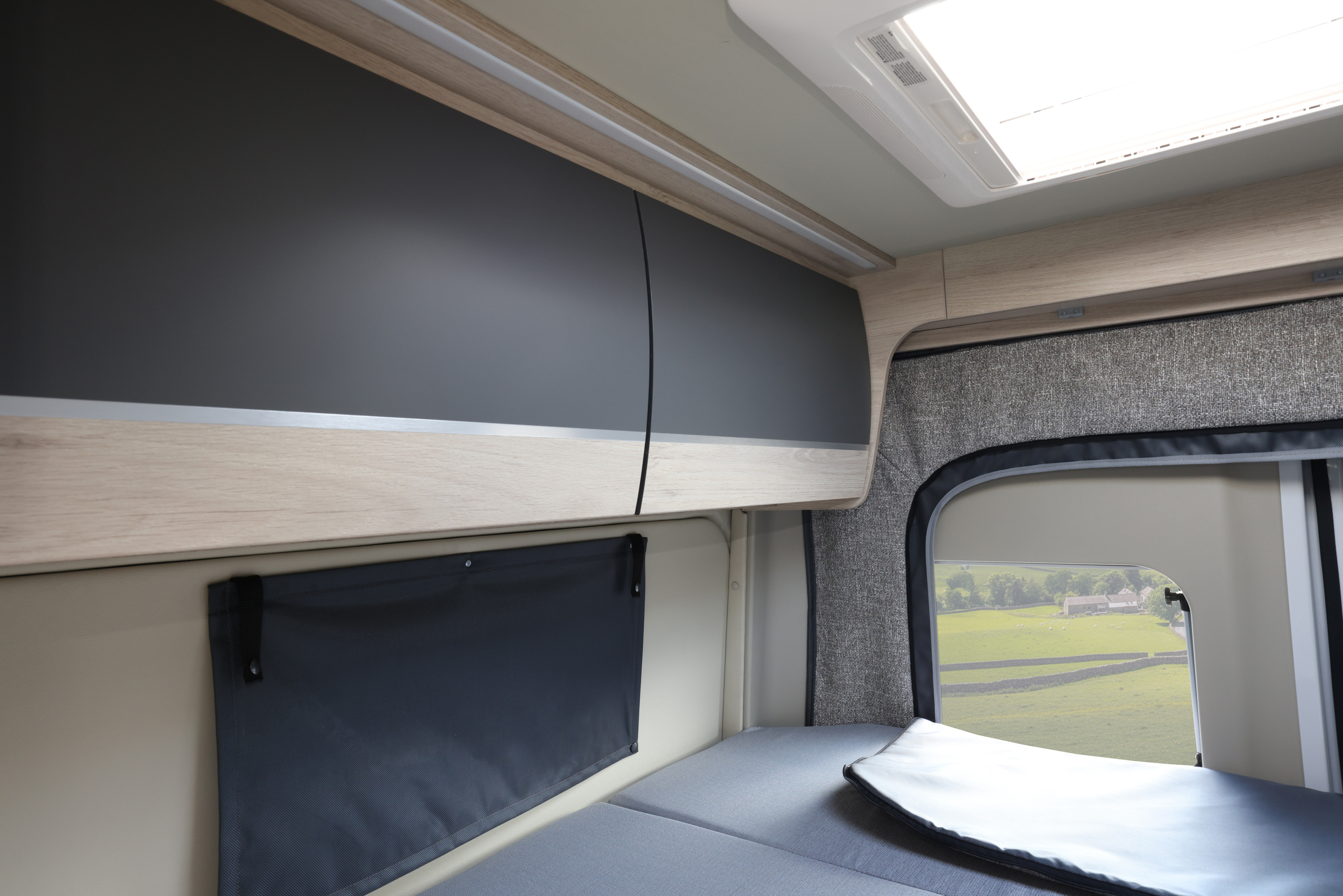 Inside a modern **RV rental**: gray overhead cabinets, strip lighting, comfortable bed, and a window revealing a serene countryside.. AutoTrail Expedition 68 Pop Top 2026