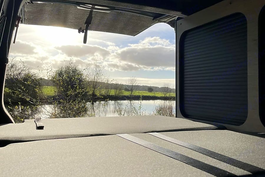 Wake up to nature's beauty in your RV rental! Cozy bed facing a tranquil river, lush fields, and bright sky.. Volkswagen summer 2024