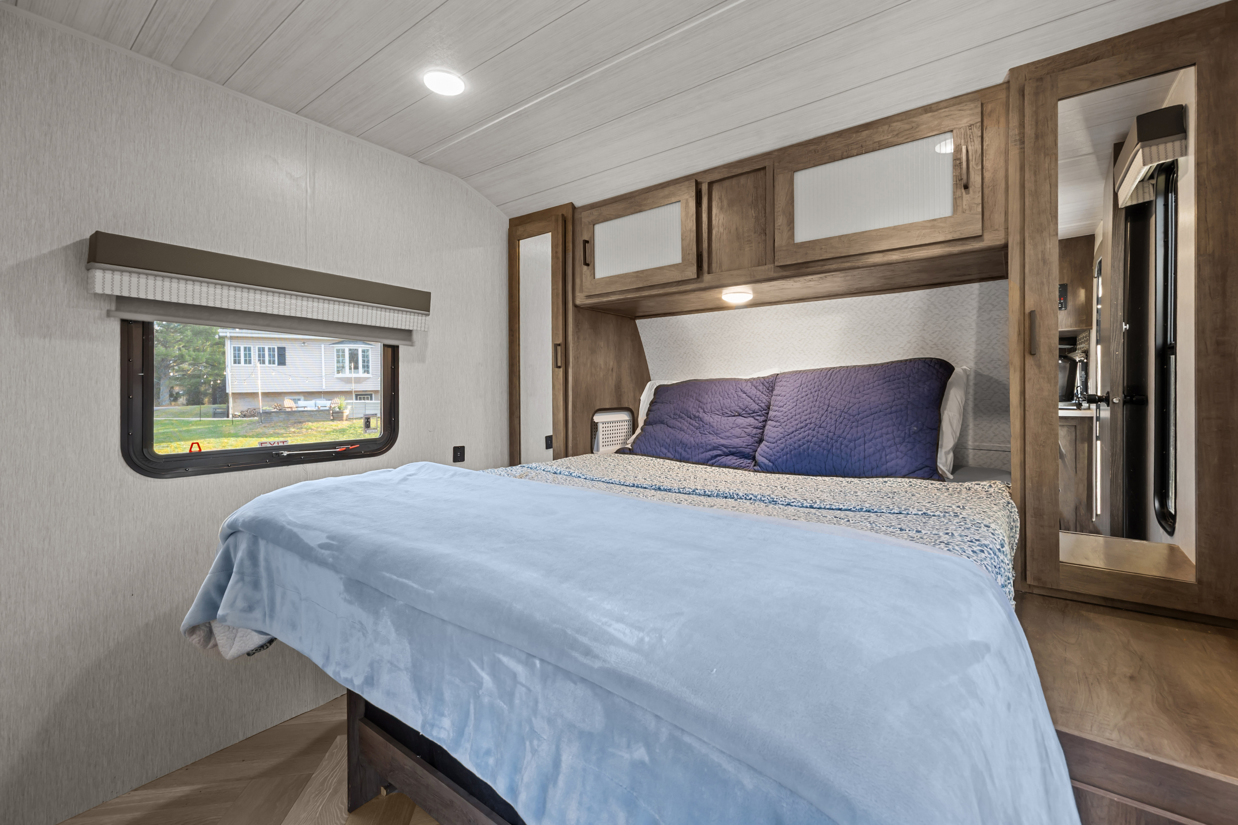 This RV rental bedroom offers a comfortable bed with blue blankets, wooden overhead cabinets, and a window to nature.. Forest River Wildwood 2022