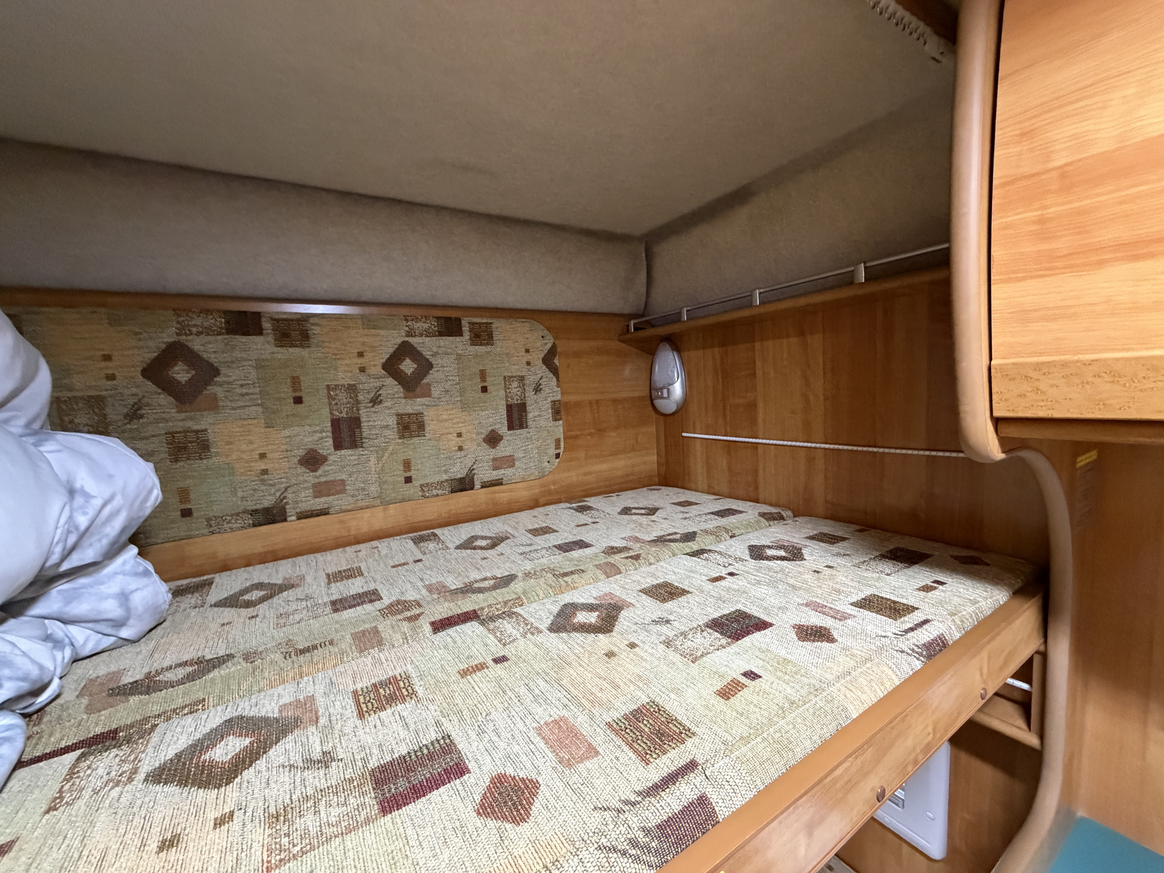 Inside an RV rental: a cozy bunk bed with patterned mattress, wooden interior, and a small reading light. White bedding visible.. Toyota Camroad 2005