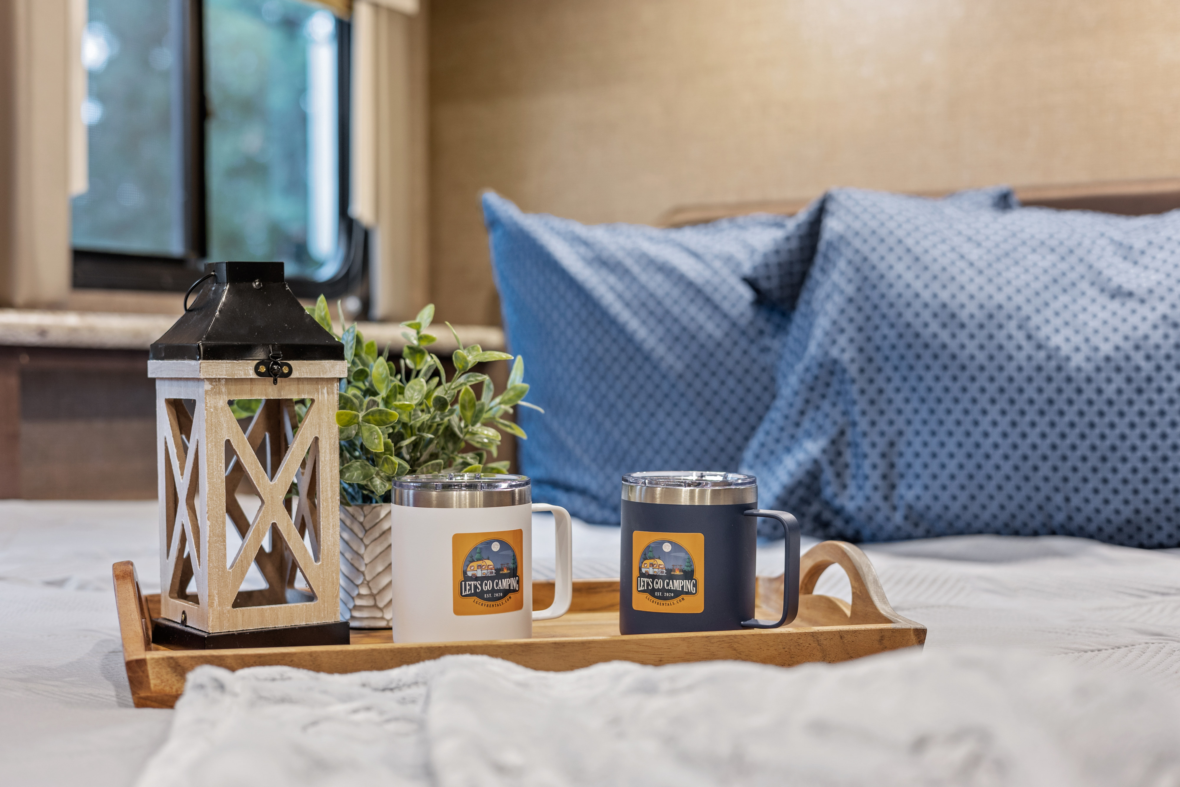 A cozy RV rental bedroom scene, with a bed tray holding 'Let's Go Camping' mugs, a lantern, and a plant.. Thor Four Winds 2017
