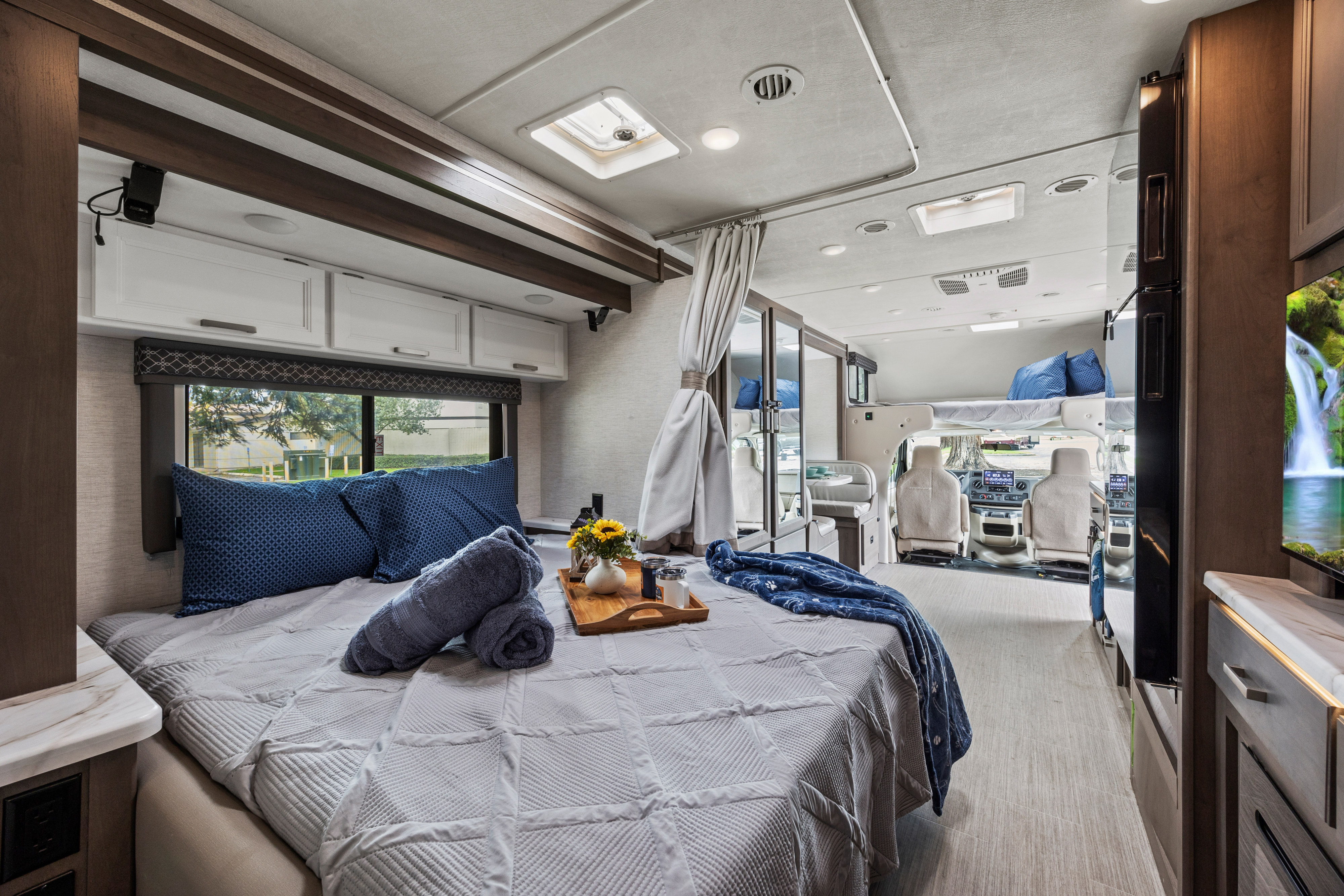 Spacious and modern RV rental interior shows a cozy bedroom with blue linens, flowing into the well-lit living and cab areas.. Thor Freedom Elite 2025