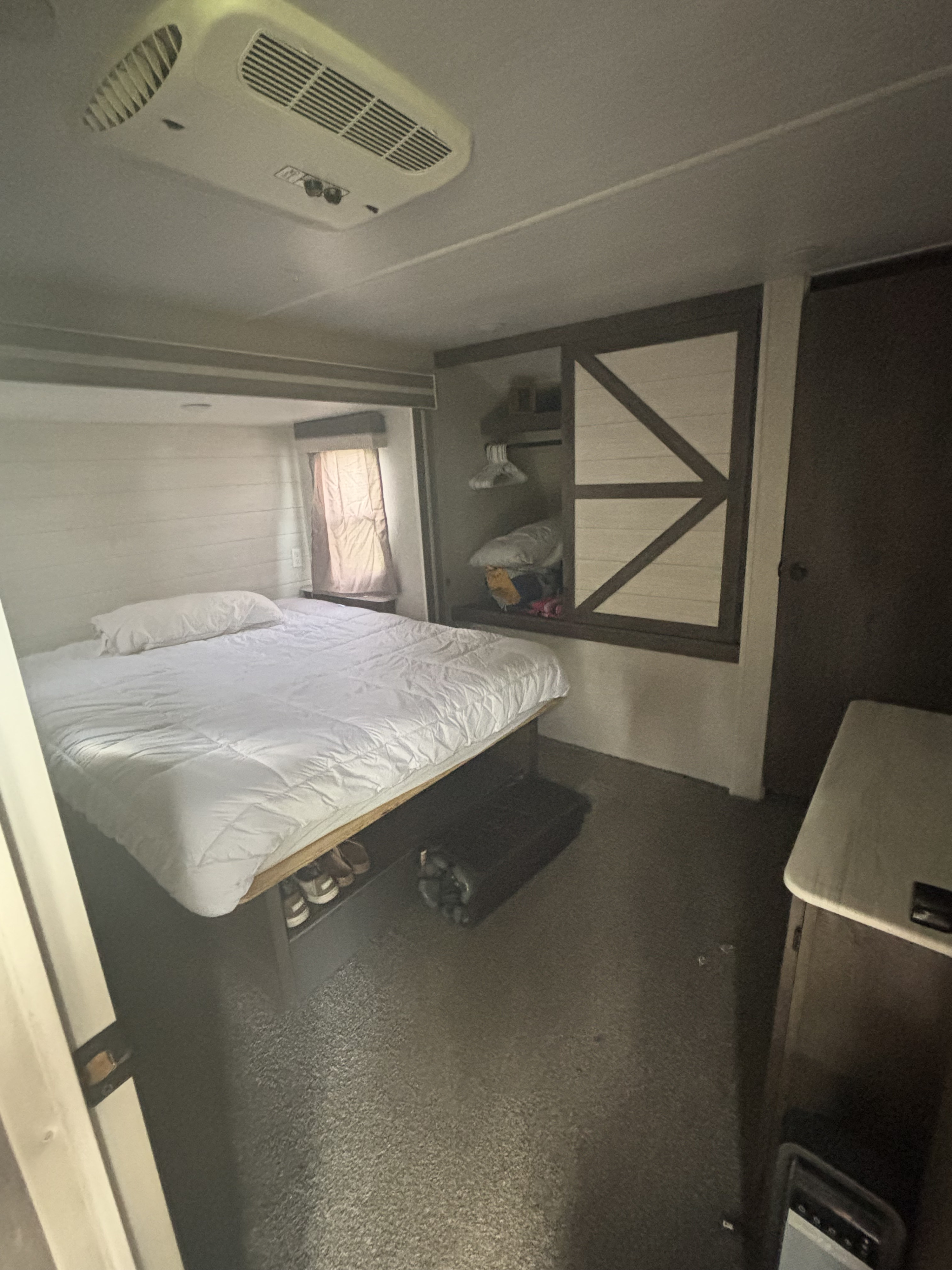 Cozy RV rental bedroom featuring a white bed, window with curtain, barn-door style closet, and AC unit. Shoes are stored under the bed.. Forest River Salem 2021