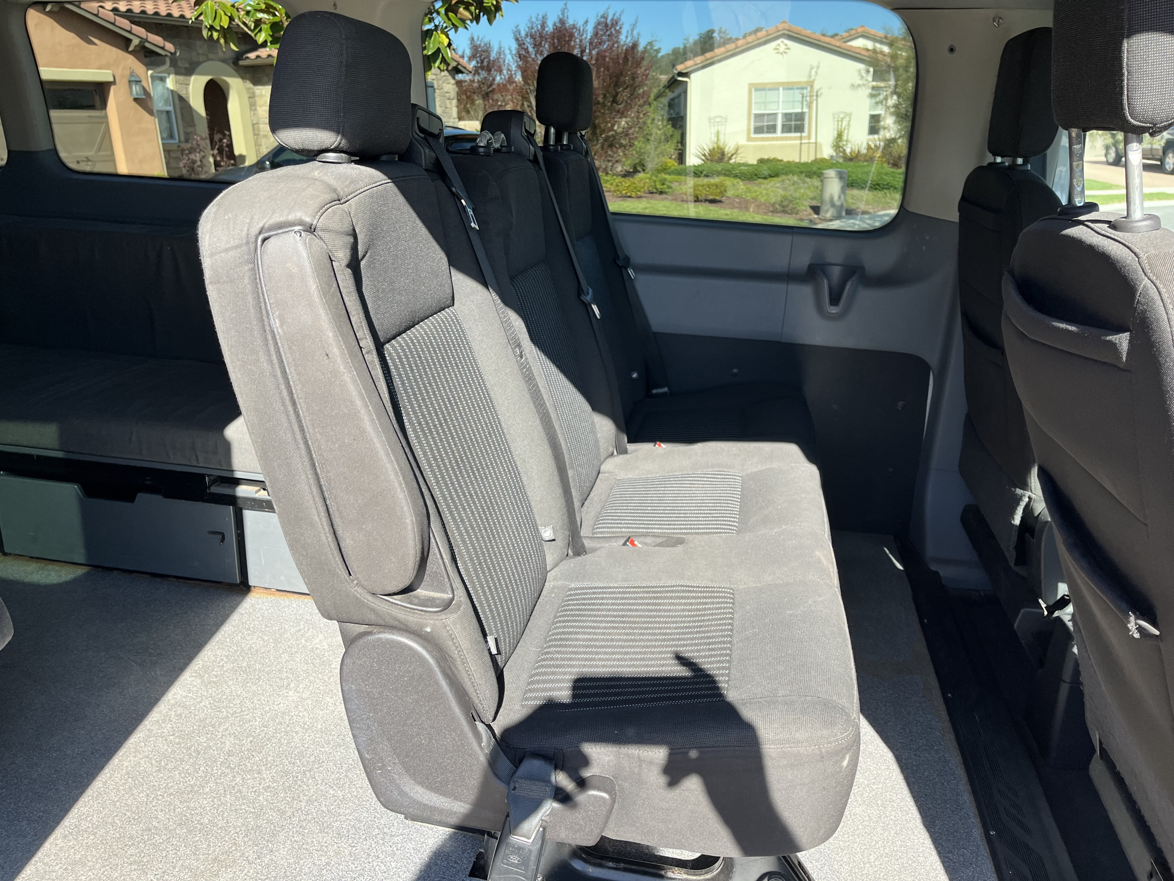 RV rental interior: black fabric passenger seats and a rear convertible bed/storage platform, with houses visible outside.. Ford Custom Transit 2017