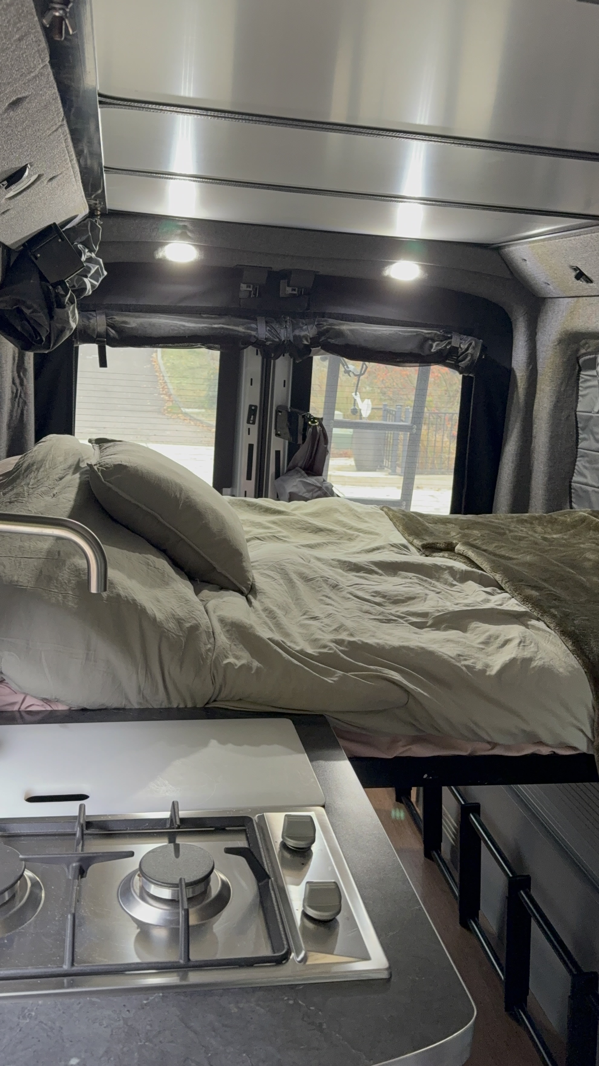 Inside an RV rental, a bed with light bedding is next to a compact kitchen with a two-burner stove and sink faucet.. ModVans ModVans Class B 2022