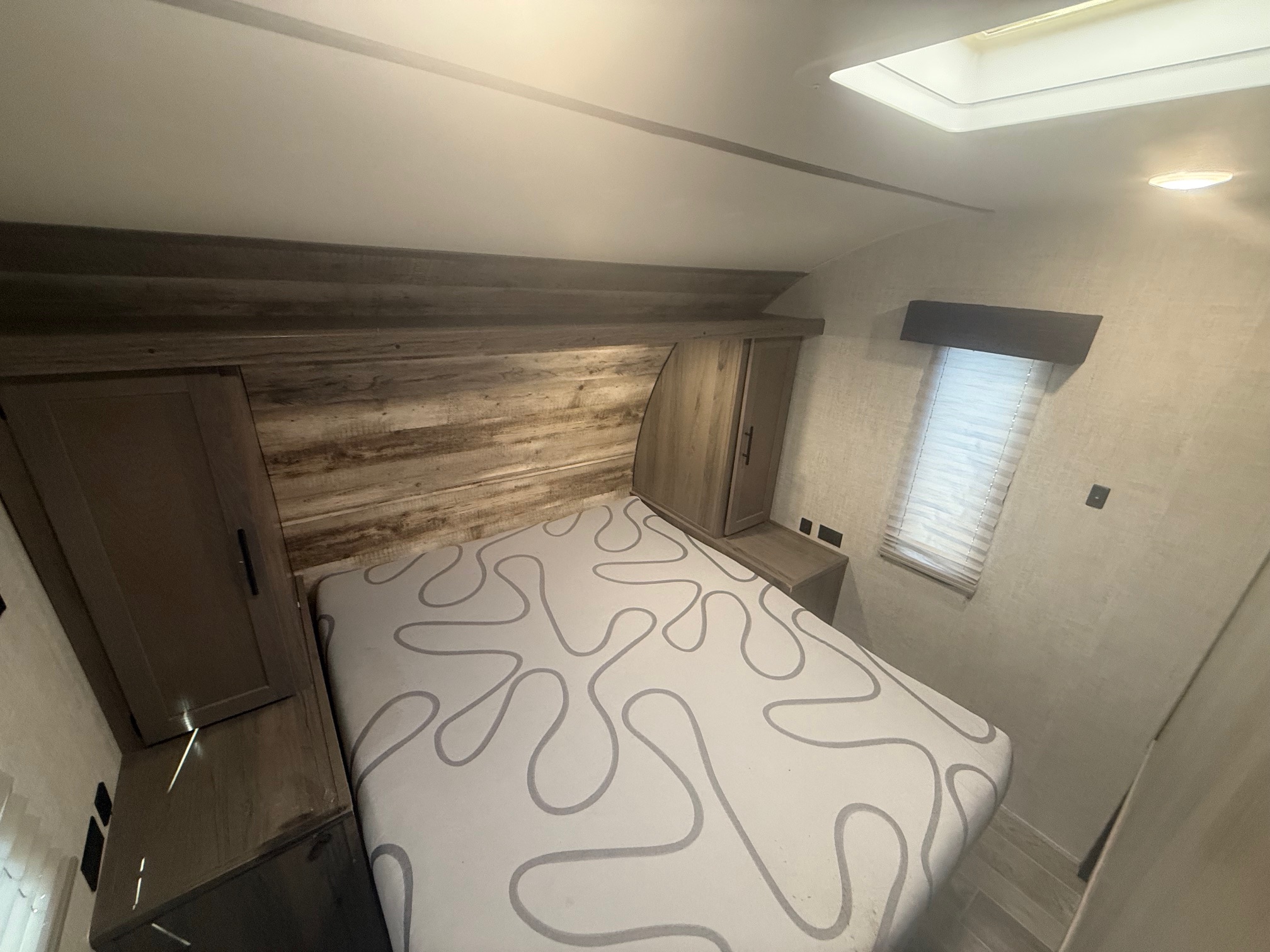An RV rental bedroom with a white bed, abstract gray pattern, wood headboard, side cabinets, window, and ceiling skylight.. Gulf Stream Kingsport 2021