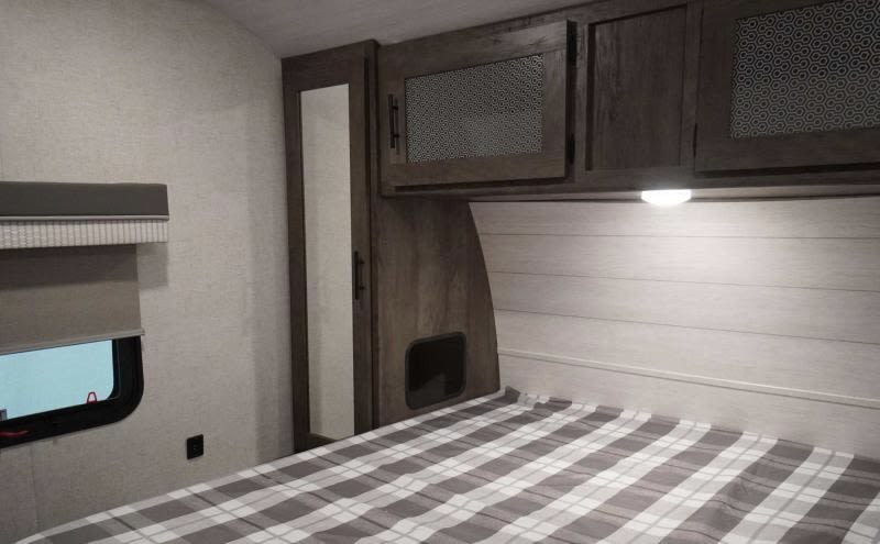 Cozy RV rental bedroom with plaid bed, overhead cabinets, mirror, and window. Perfect for your next adventure.. Forest River Wildwood 2021