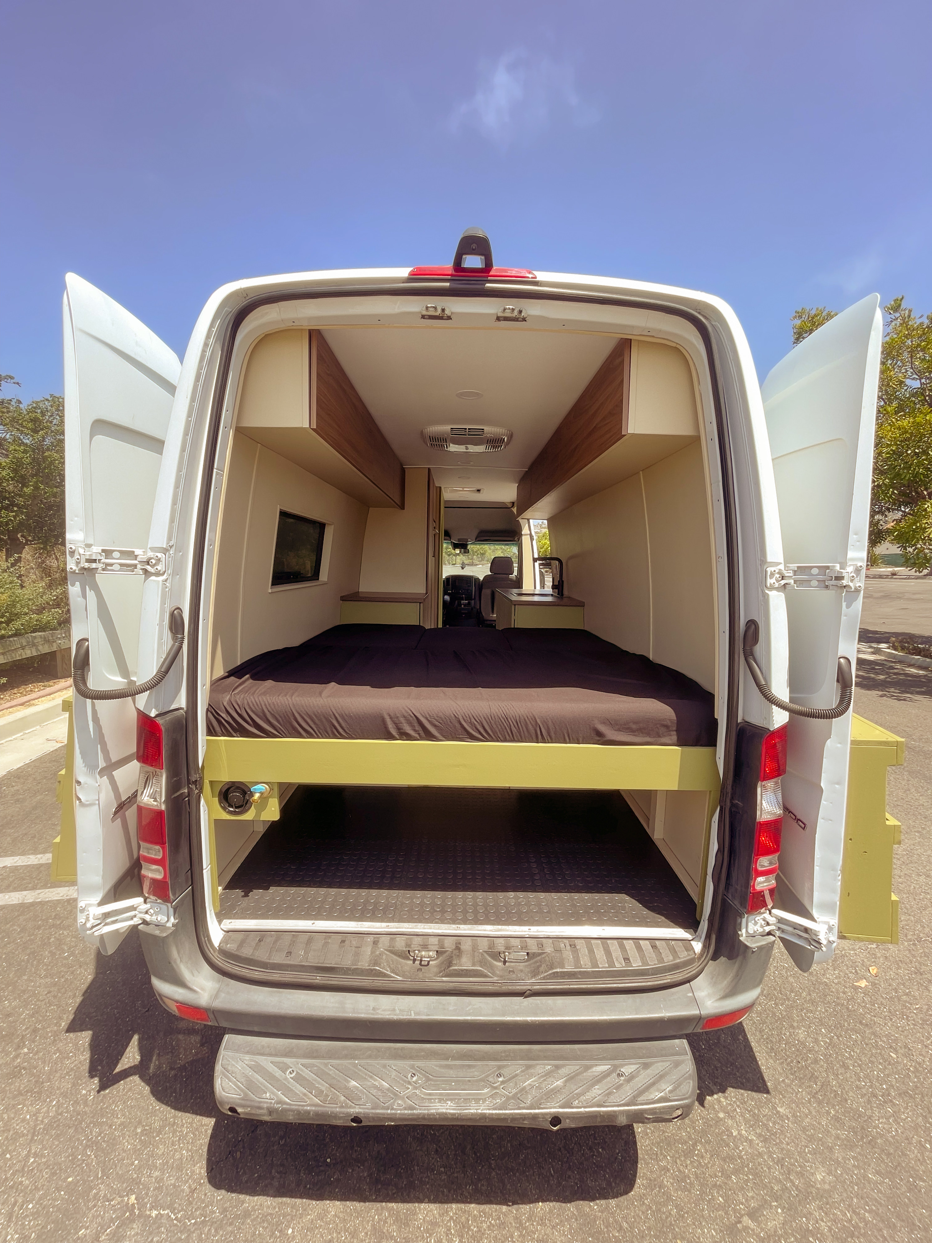 Interior of an RV rental campervan with open doors: a bed, under-bed storage, overhead compartments, and a kitchen.. Mercedes-Benz Sprinter RV Motorhome Campervan 2018