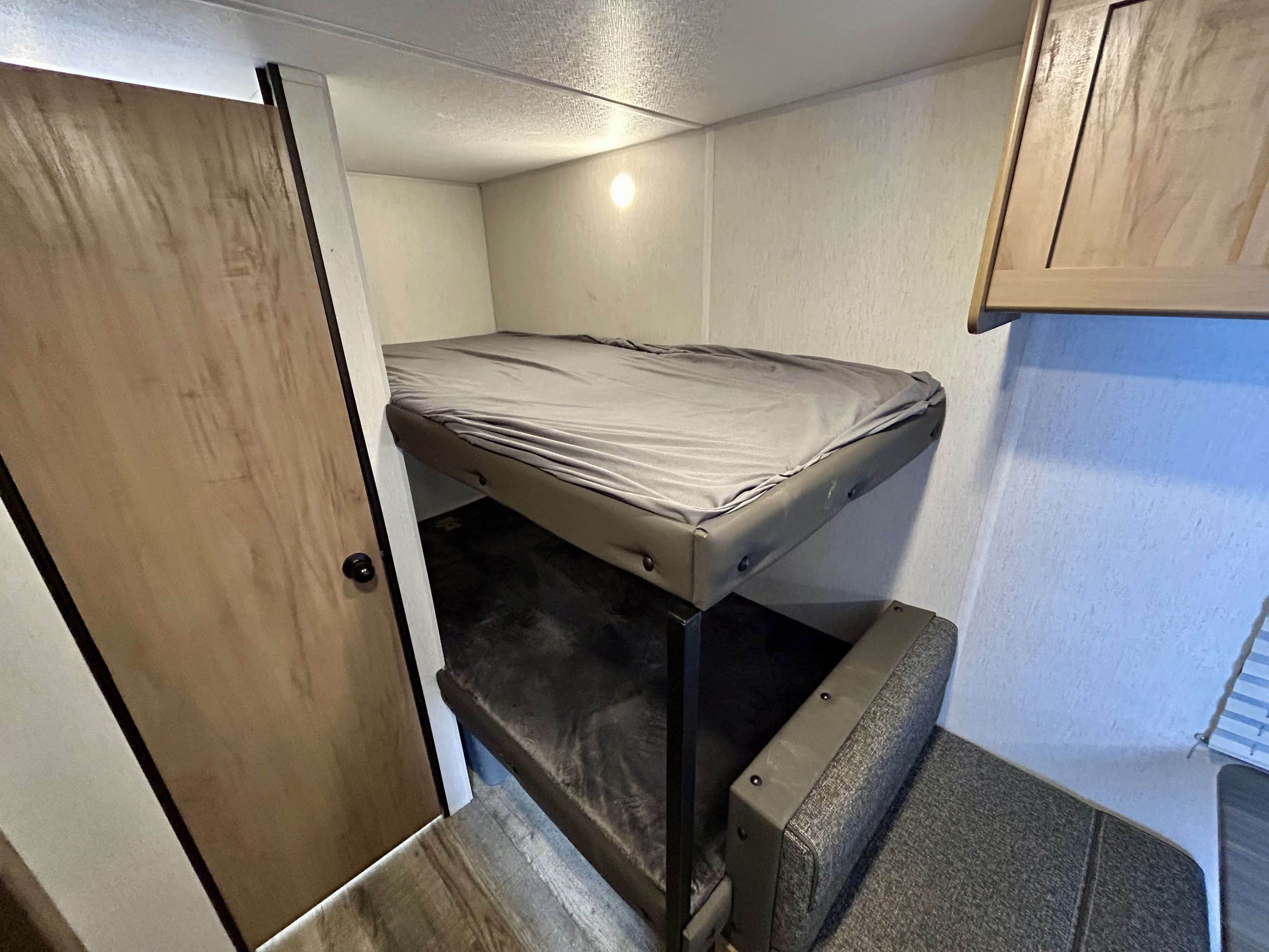 Compact bunk bed area inside an RV rental, with grey sheets, a wall light, and a light-colored wooden door visible.. Forest River Cherokee Wolf Den 2024