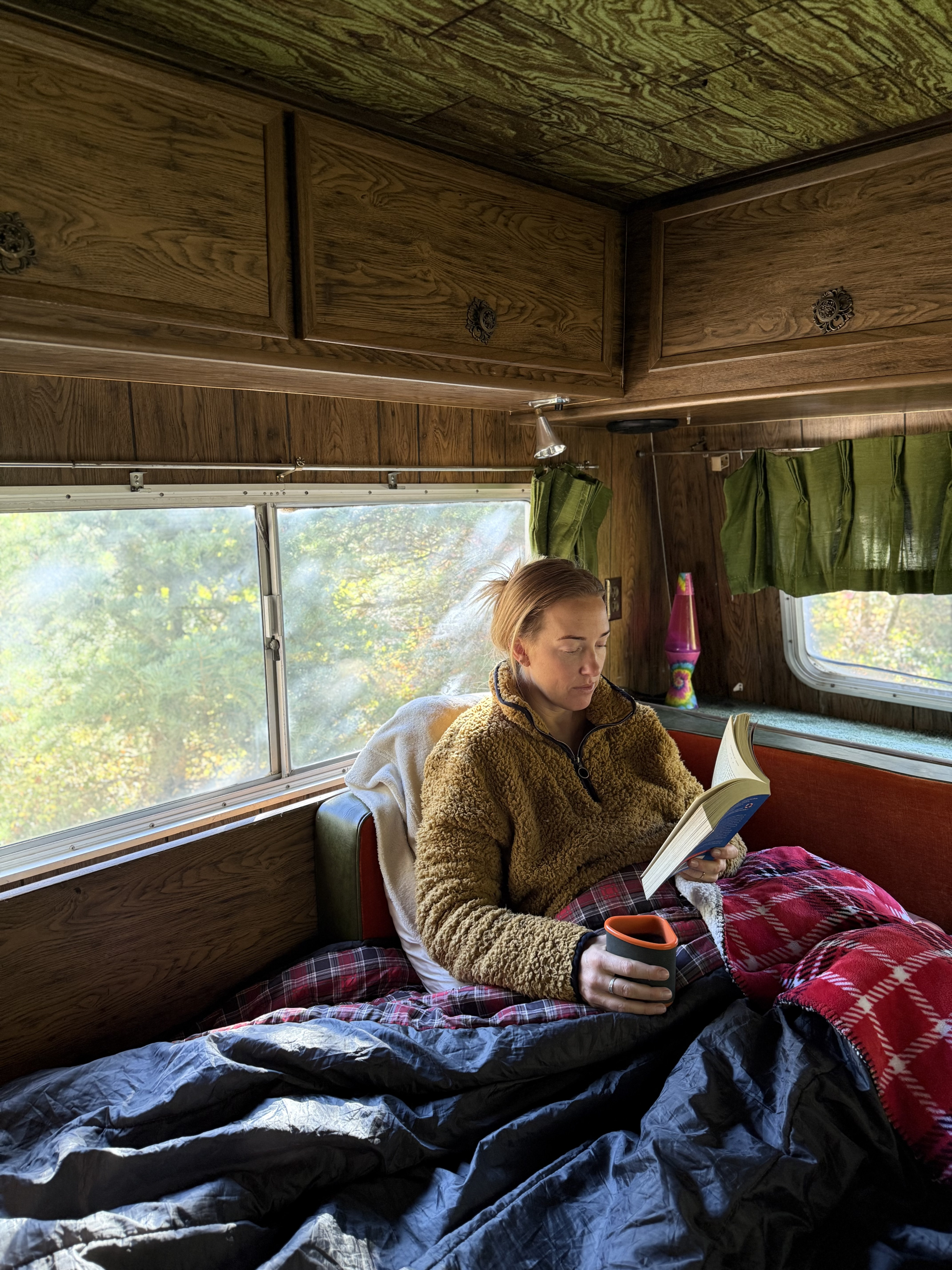 Cozy inside an RV rental: a woman reads a book & sips from a mug, surrounded by vintage wood paneling & nature outside.. International 610A 1973