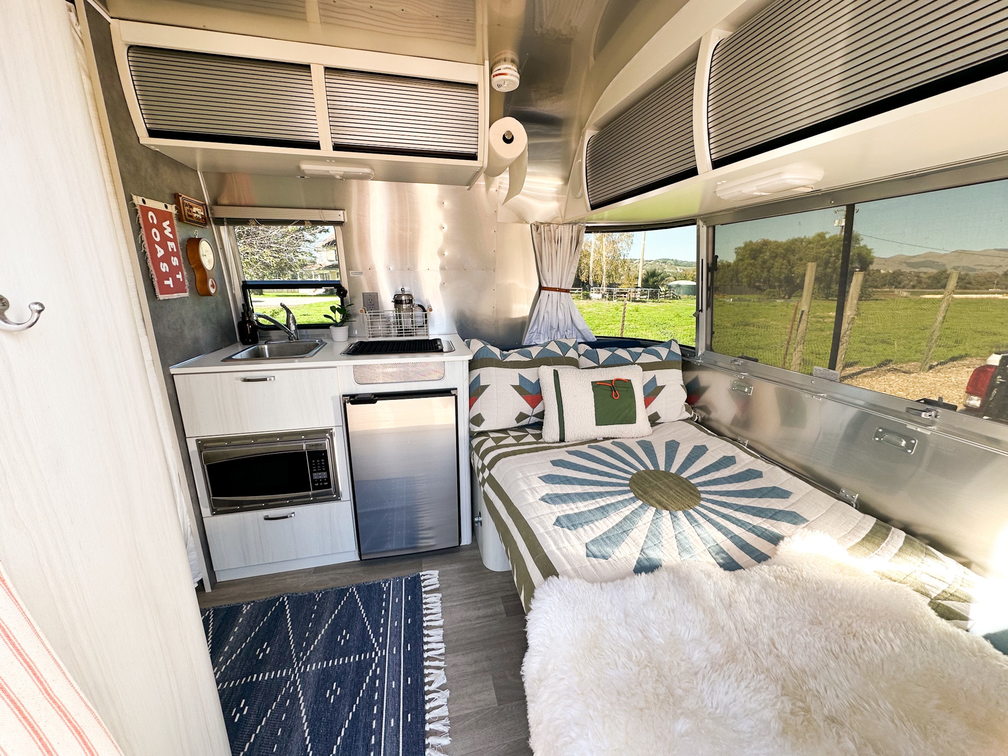 Bright RV rental interior showcasing a compact kitchenette, a bed with a colorful quilt, and a scenic outdoor view.. Airstream Bambi 2020