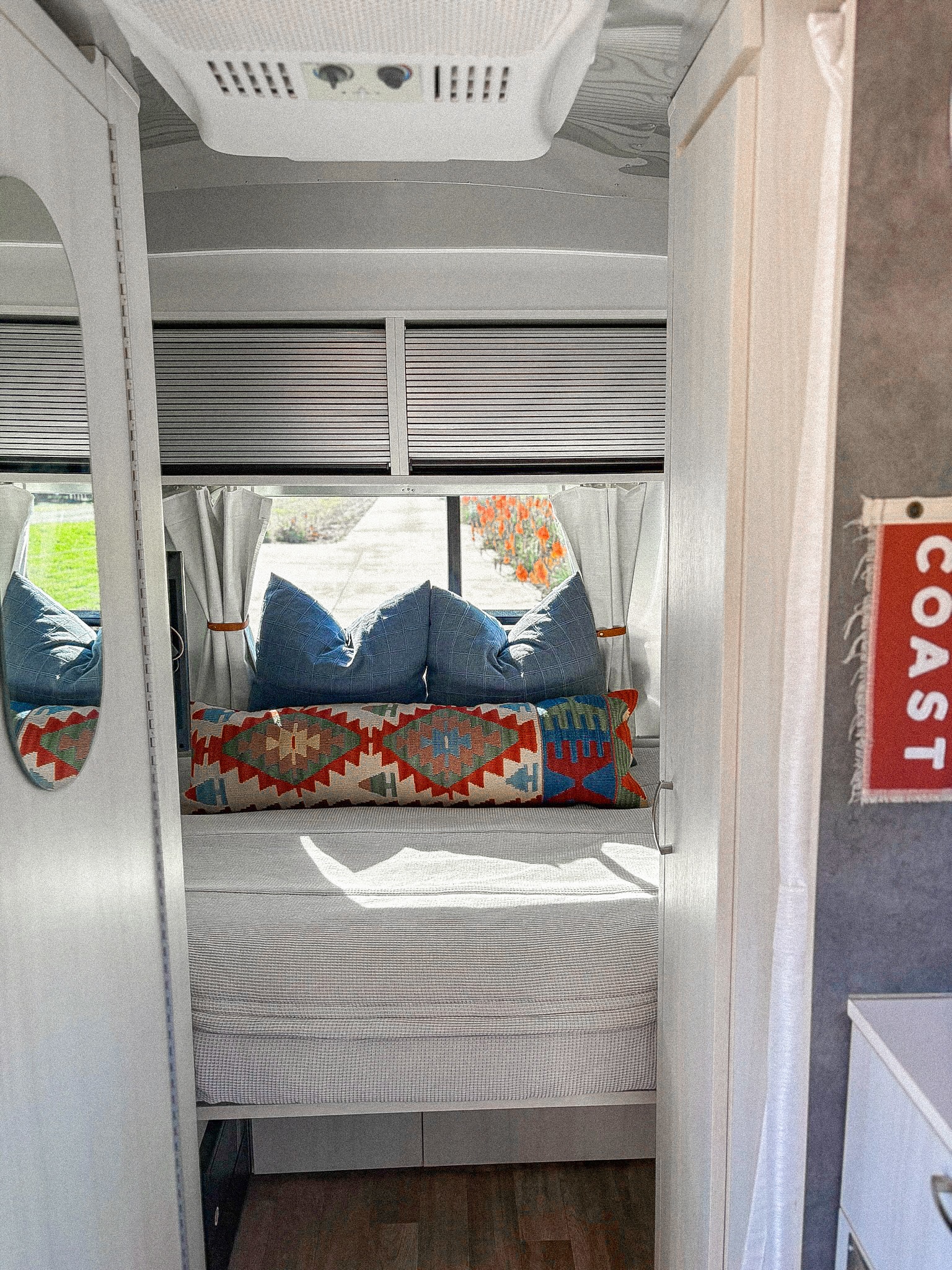 Cozy rv rental interior, showing a bed with blue and patterned pillows, window blinds, and an AC unit for comfortable travel.. Airstream Bambi 2020