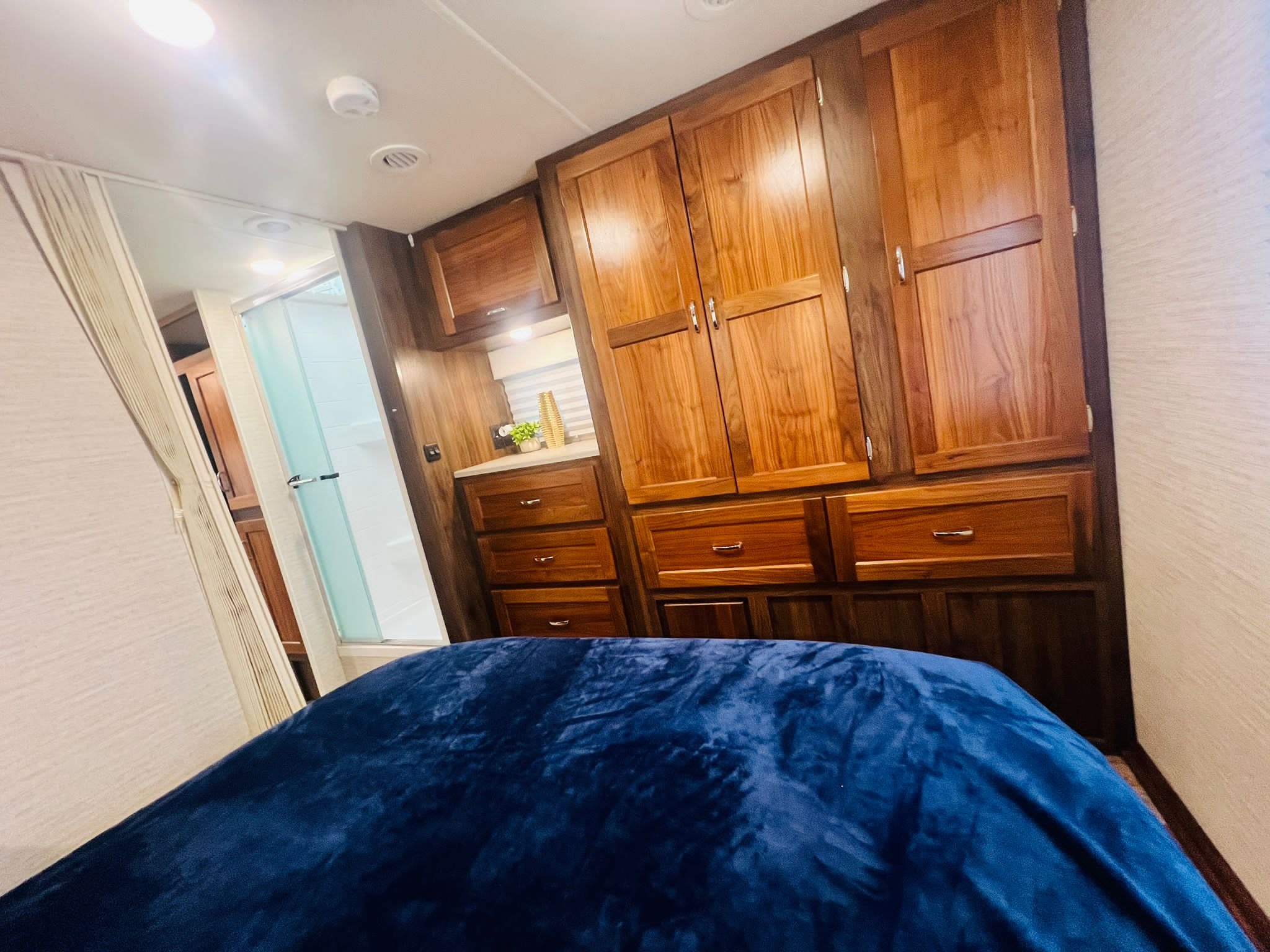 This RV rental interior features a cozy bedroom with a blue blanket, extensive wood cabinets, and a compact bathroom with a shower.. Winnebago Outlook 2020