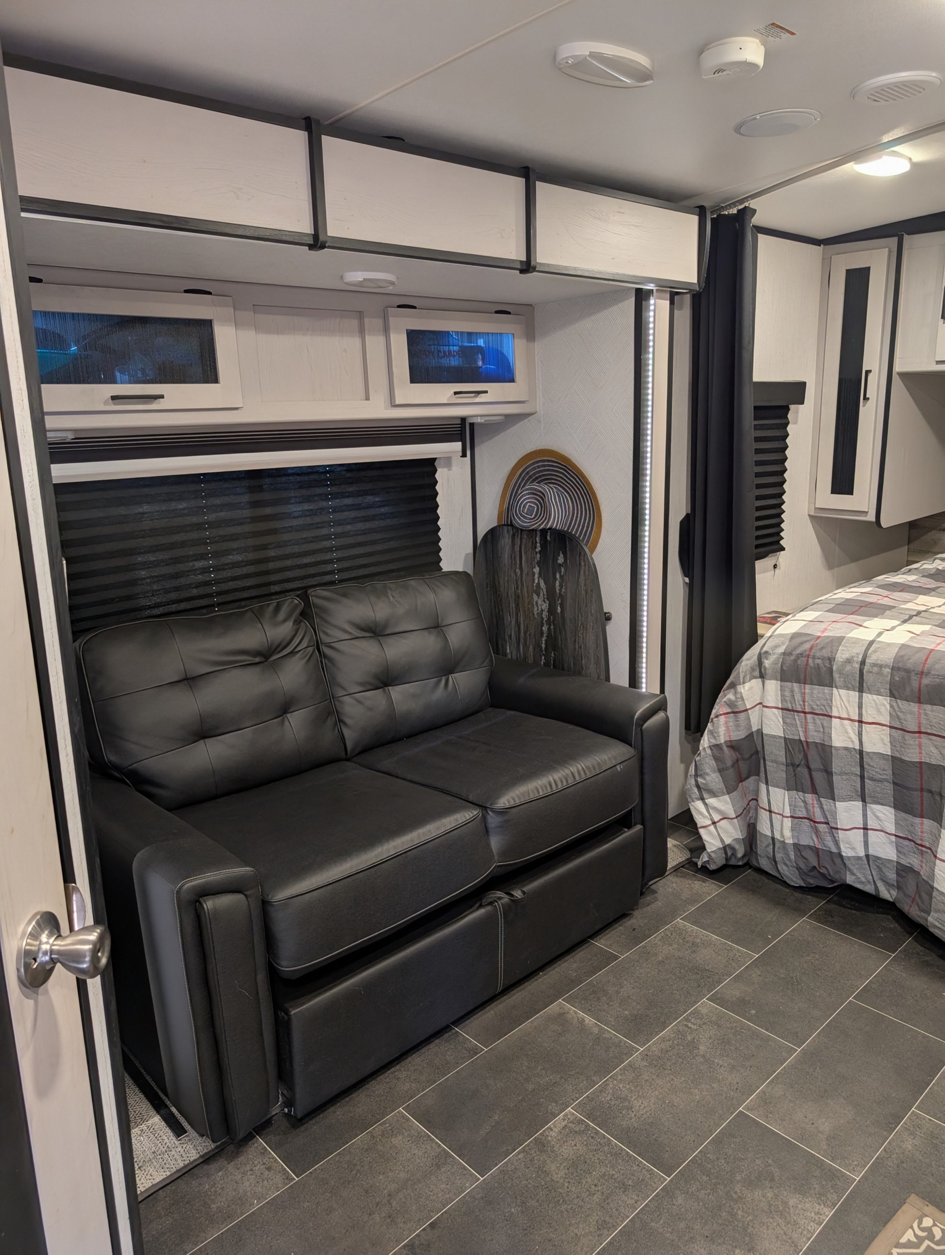 An RV rental's inviting interior showing a black leatherette sofa, overhead storage, and a comfy plaid bed.. Heartland RVs Mallard 2022
