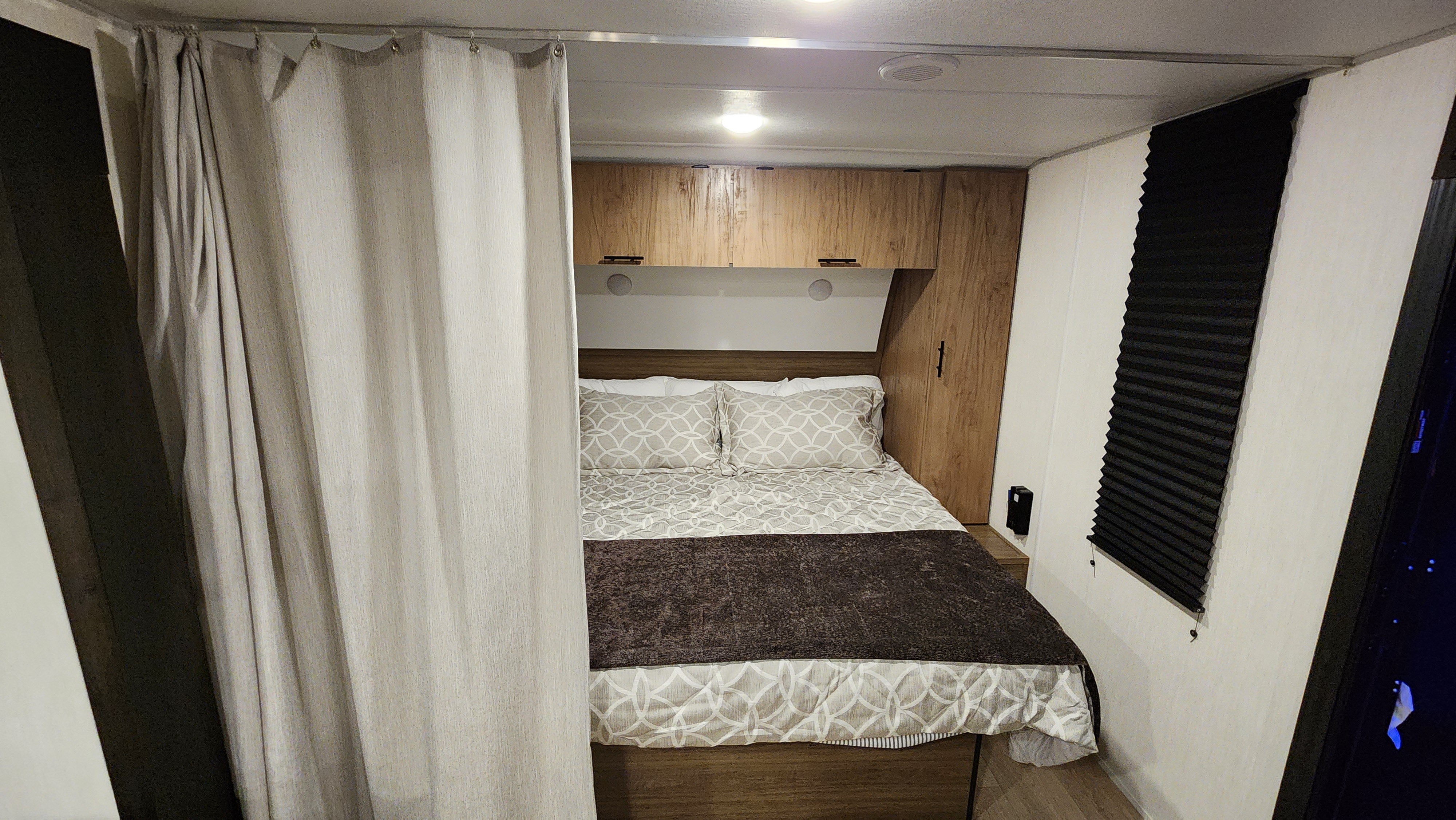 Cozy RV rental bedroom featuring a bed with patterned bedding, privacy curtains, wood-look cabinets, and a pleated window blind.. Forest River Cherokee Grey Wolf 2025
