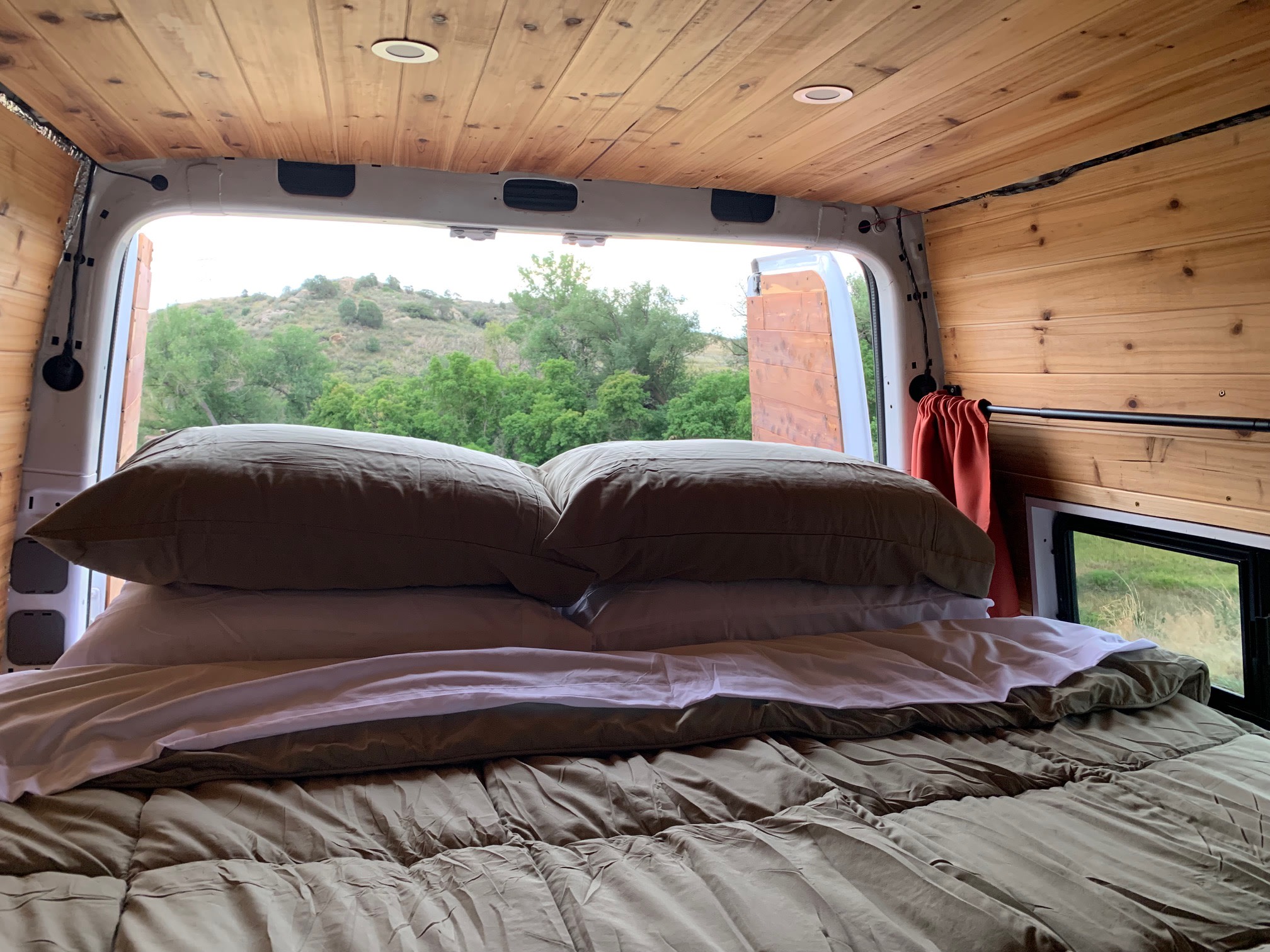 Cozy rv rental bed inside a custom van, featuring wooden walls/ceiling and a scenic view of green hills and trees.. Mercedes Sprinter 2011