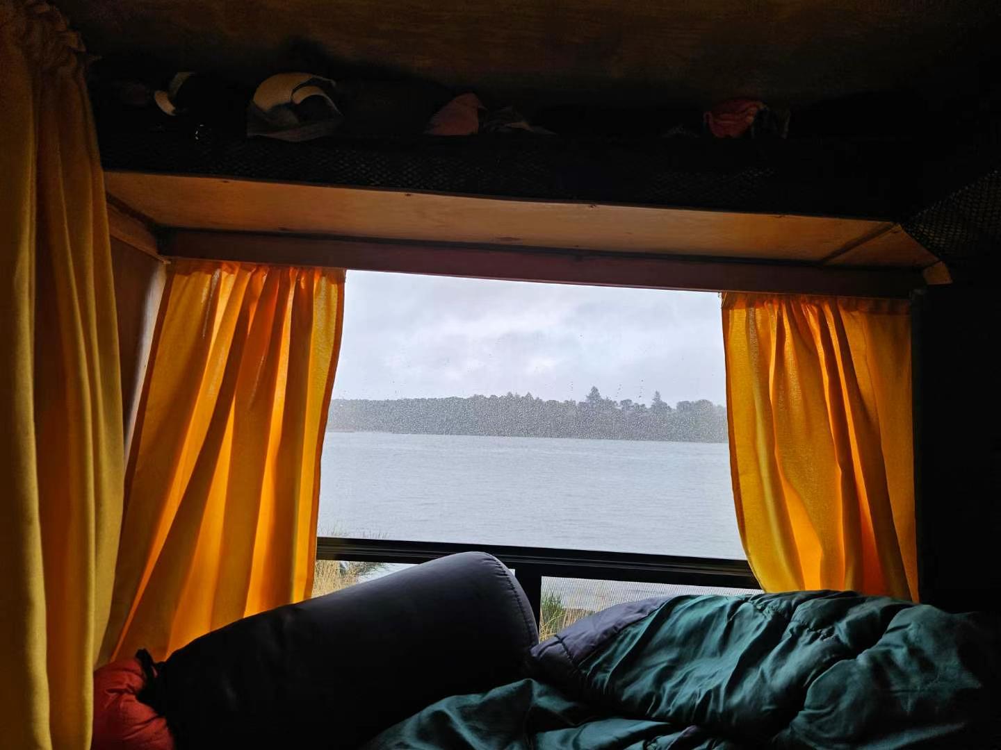 Enjoying a serene lake view from a cozy RV rental bedroom. Yellow curtains frame a cloudy sky over water and distant trees.. Chevrolet Express 2022