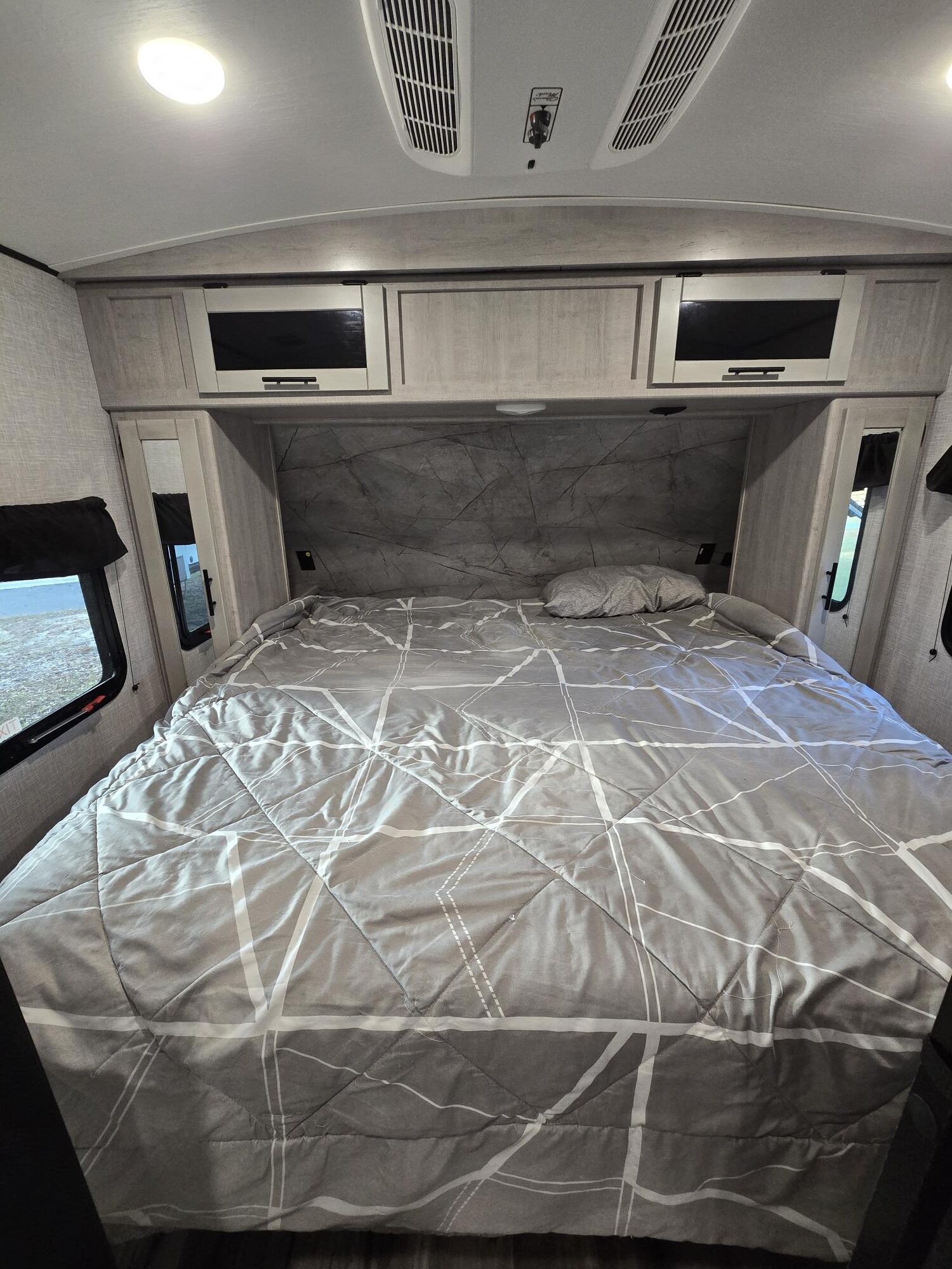 Modern RV rental bedroom with a large bed, grey geometric comforter, overhead storage, AC vent, and side windows.. Crossroads RV Sunset Trail 2023