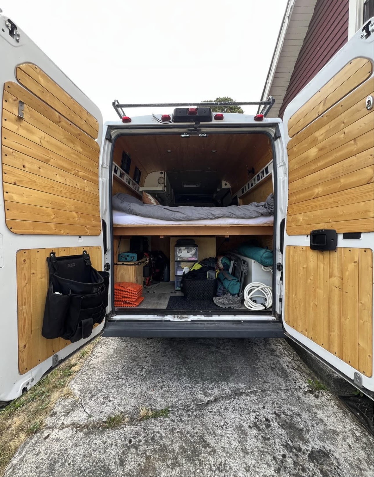 Open back of an RV rental camper van showing custom wood interior, a bed with grey bedding, and organized storage below.. Dodge Ram 2500 ProMaster 2015
