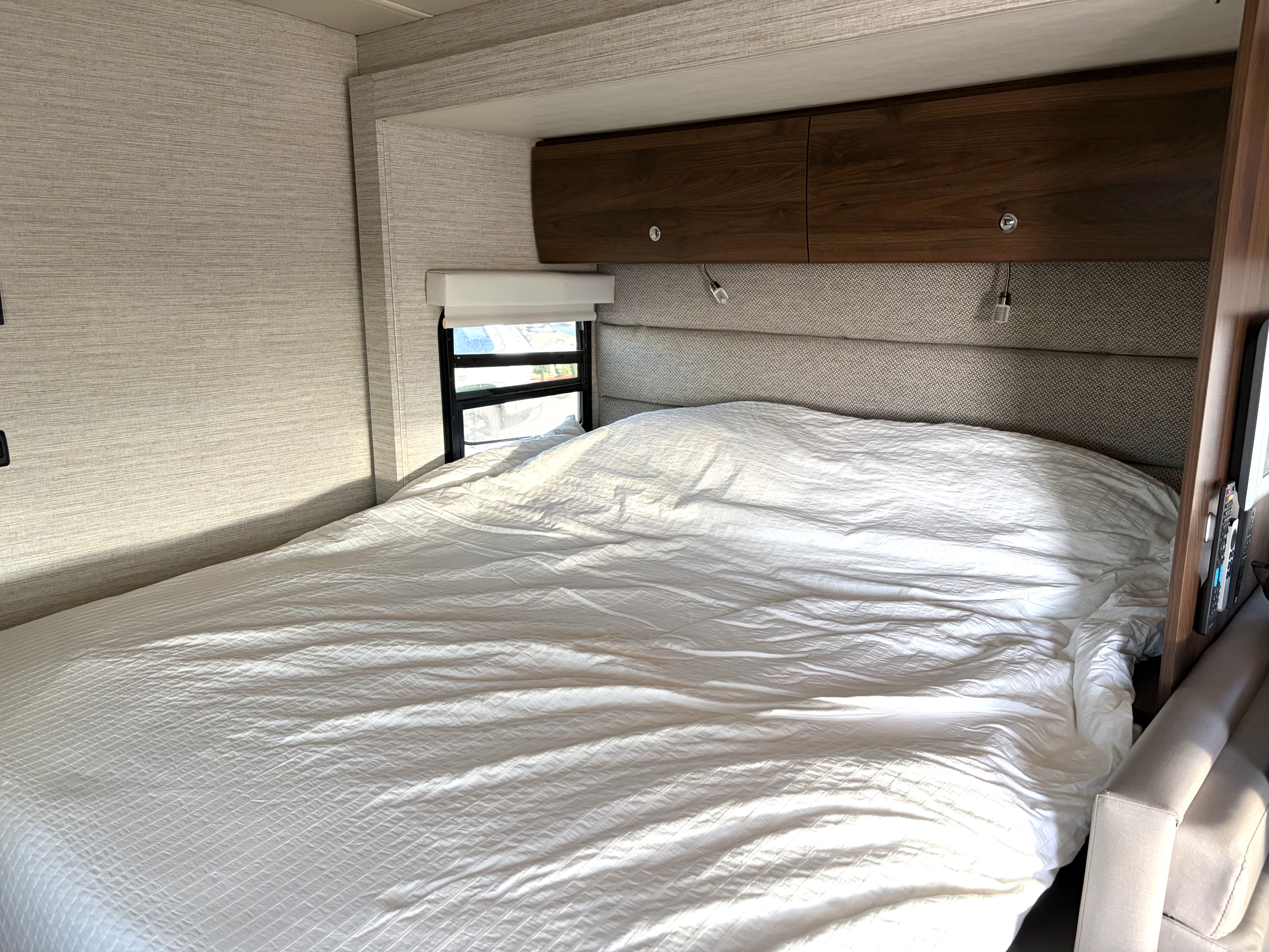 Cozy RV rental bedroom with a white textured bed, overhead storage, reading lights, and a window by the headboard.. Winnebago View 2022