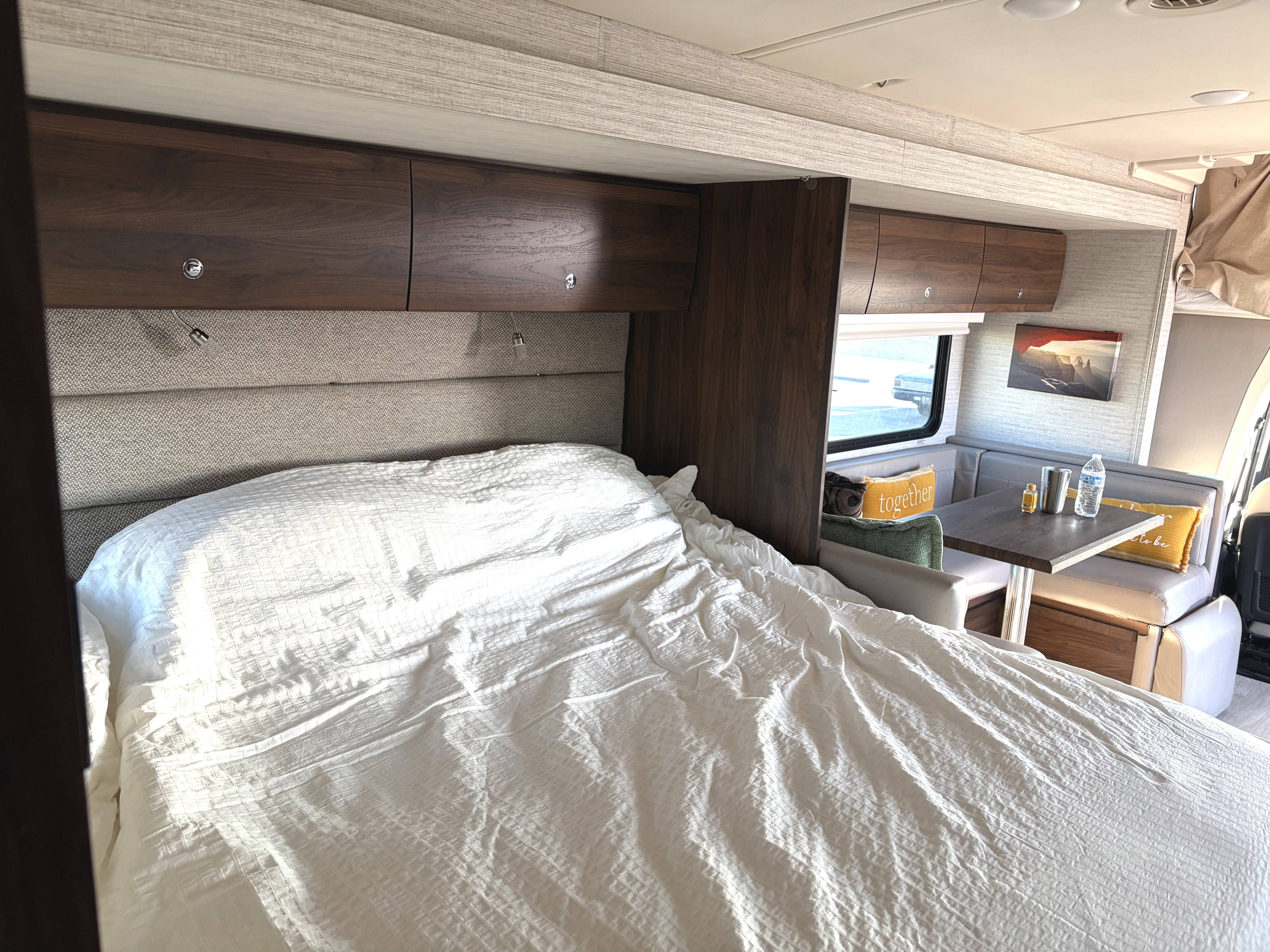 RV rental interior featuring a comfy bed with overhead storage and a dining area with a table and bench seating.. Winnebago View 2022