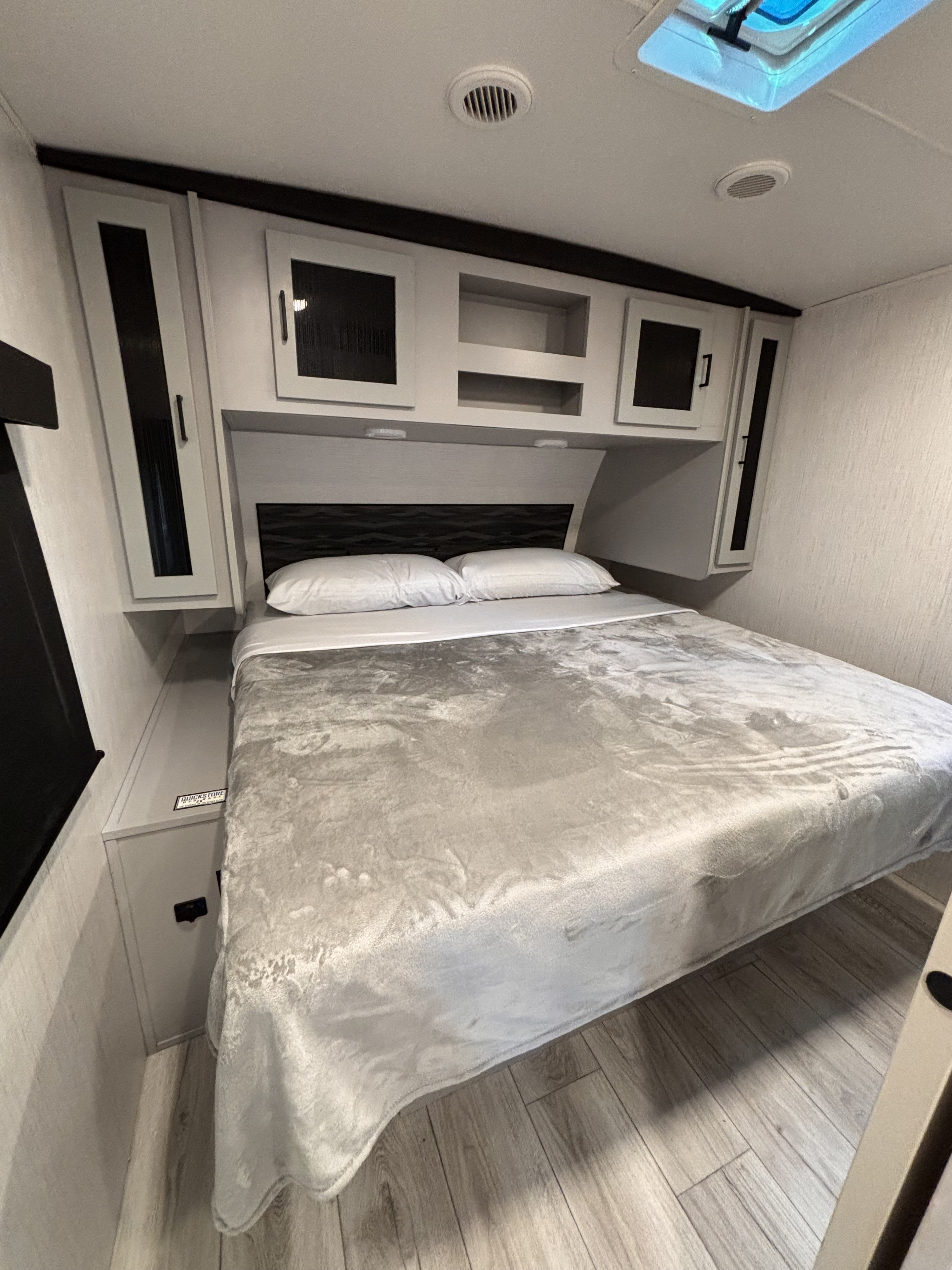 Cozy RV rental bedroom: a bed with white pillows and grey blanket, overhead cabinets, a dark headboard, and wood-look floor.. Heartland RVs North Trail 2022