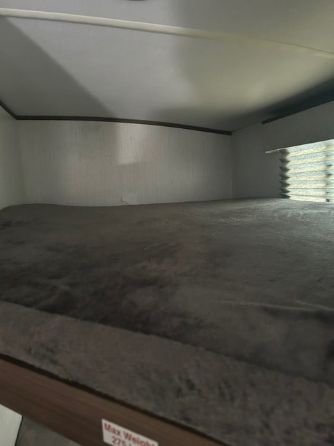 Empty bunk bed in an RV rental, featuring a dark grey blanket, white walls, and a window blind. Max weight 275 lbs.. Cruiser RV MPG 2023