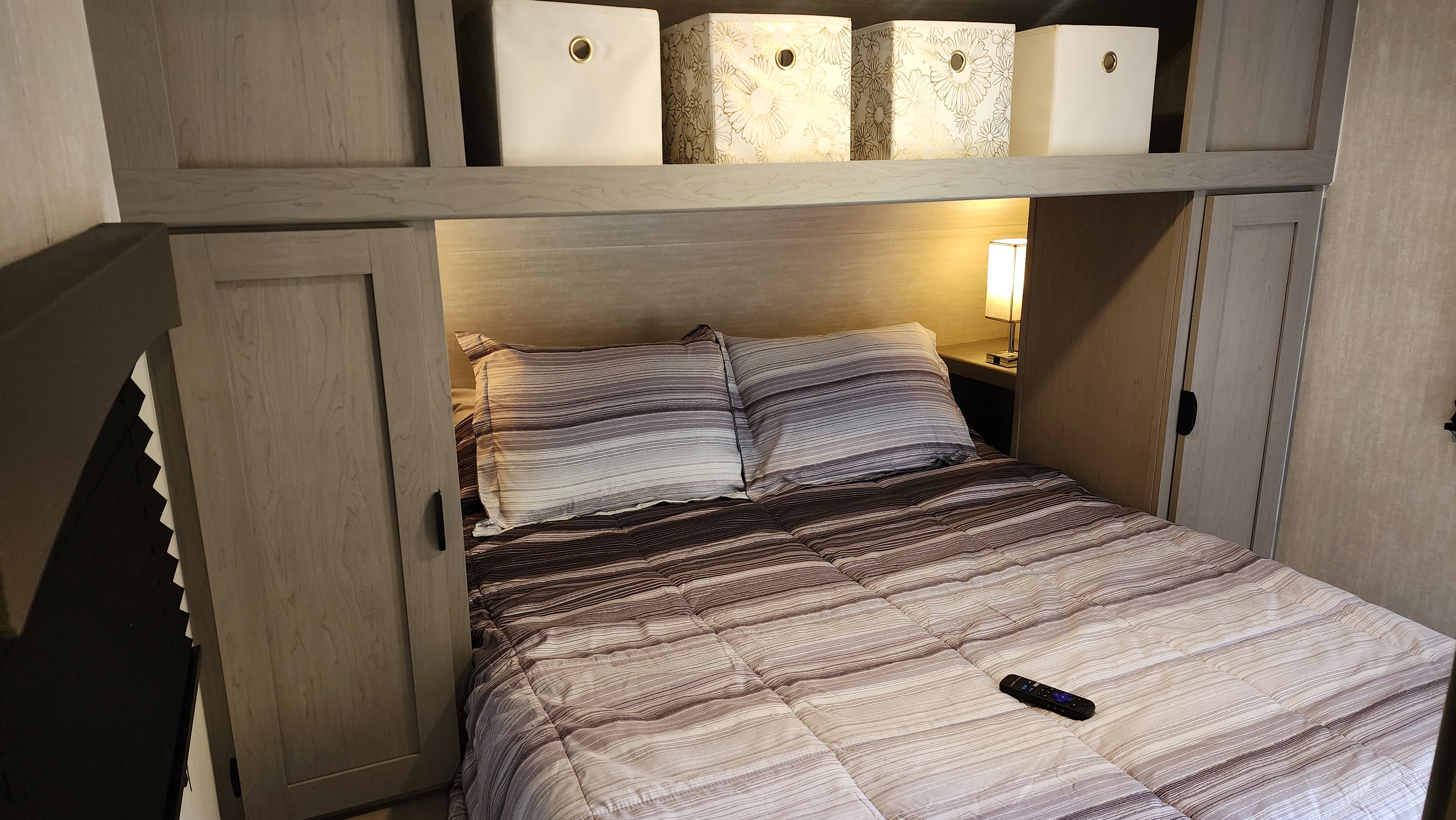 Relax in this RV rental bedroom with striped bedding, wood-toned cabinets, overhead storage bins, and a bedside lamp.. Forest River Viking 6K 2025