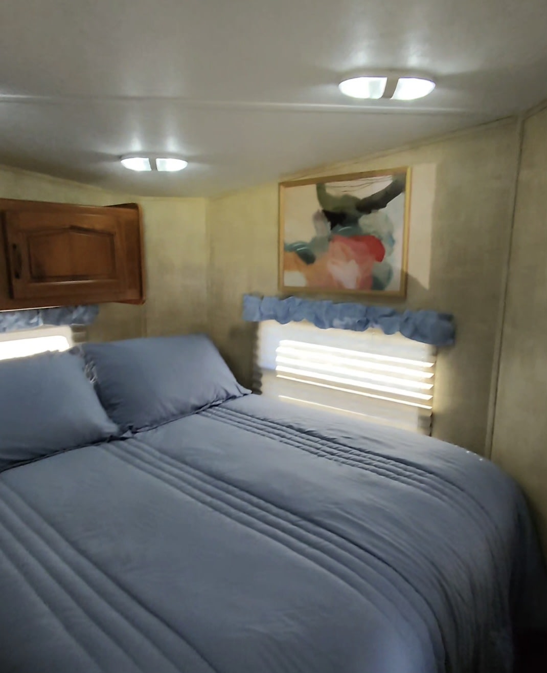 An RV rental bedroom with a blue-sheeted bed, bright overhead lights, wood cabinet, and abstract art for a cozy stay.. Keystone RV Raptor Toy Hauler 2012
