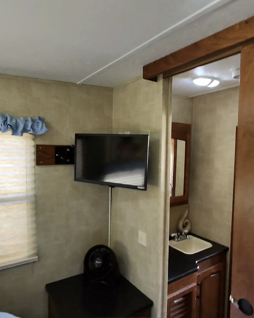 Inside an RV rental: a wall-mounted TV & fan near a window, with a bathroom sink visible through the open doorway.. Keystone RV Raptor Toy Hauler 2012