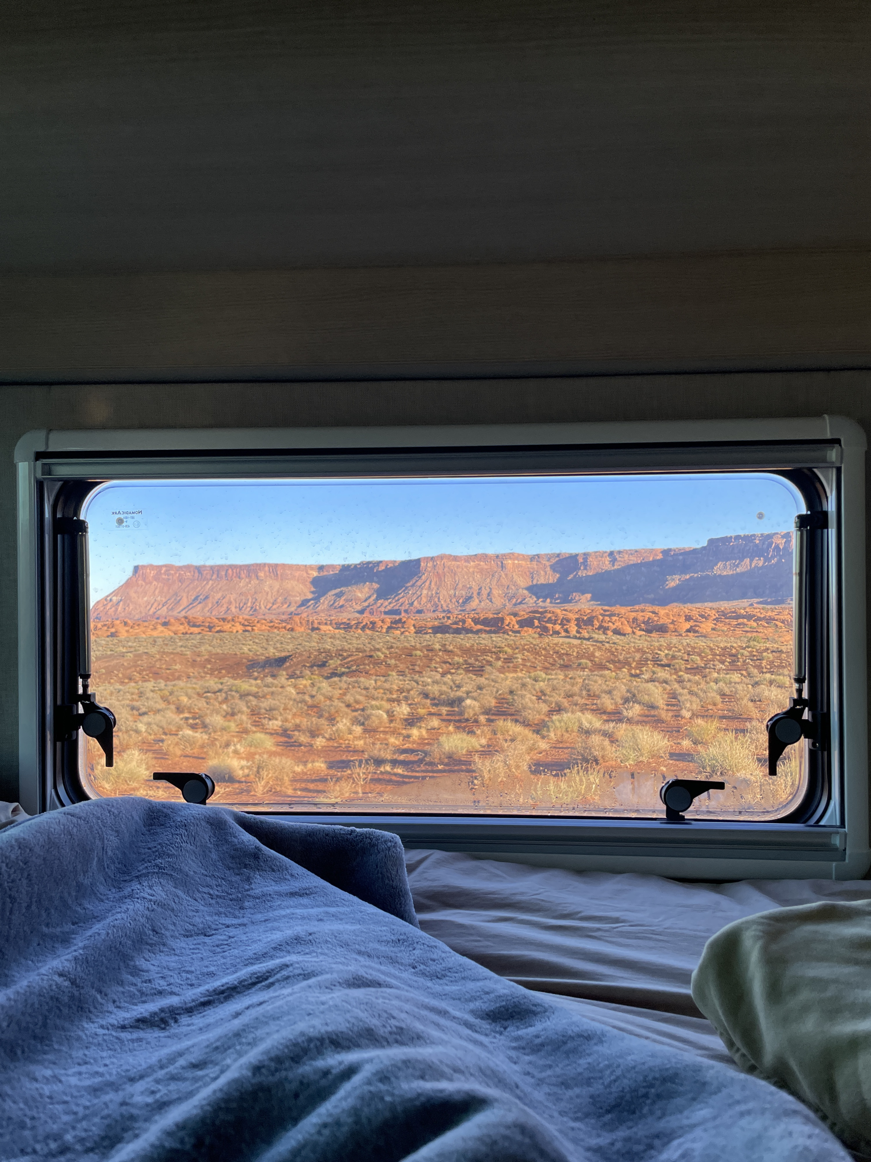 Cozy RV rental bed with a view of red rock desert mesas and scrubland through the window, offering a serene nature escape.. Custom Camper 2025
