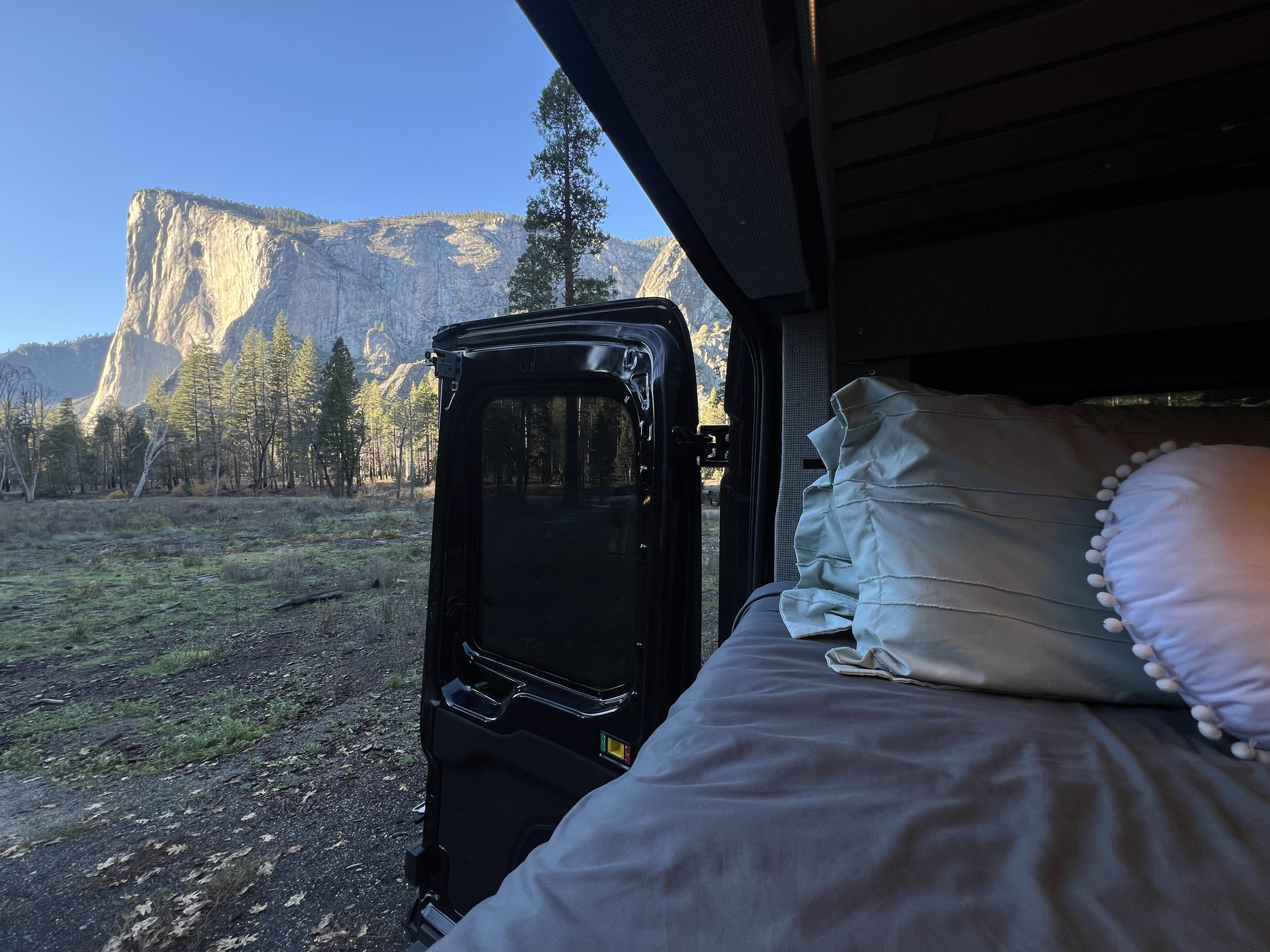 Cozy RV rental bed with an open door showcasing Yosemite's iconic El Capitan & forests under a bright blue sky. Perfect van life!. Ford Transit 2023