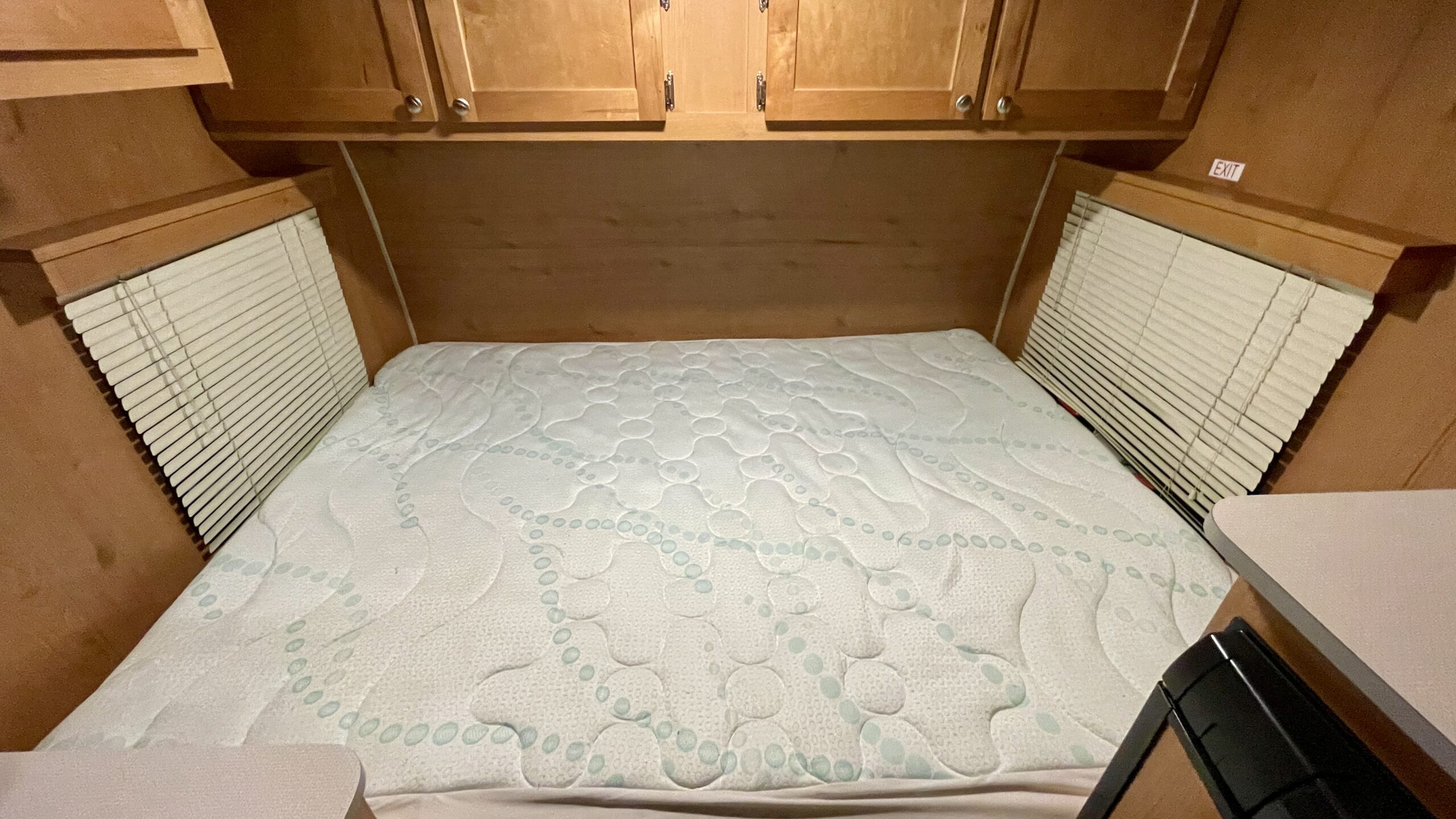 Cozy RV rental bedroom featuring a patterned mattress, wooden cabinets, and side blinds. An "EXIT" sign is visible above the right window.. Riverside Retro 2018