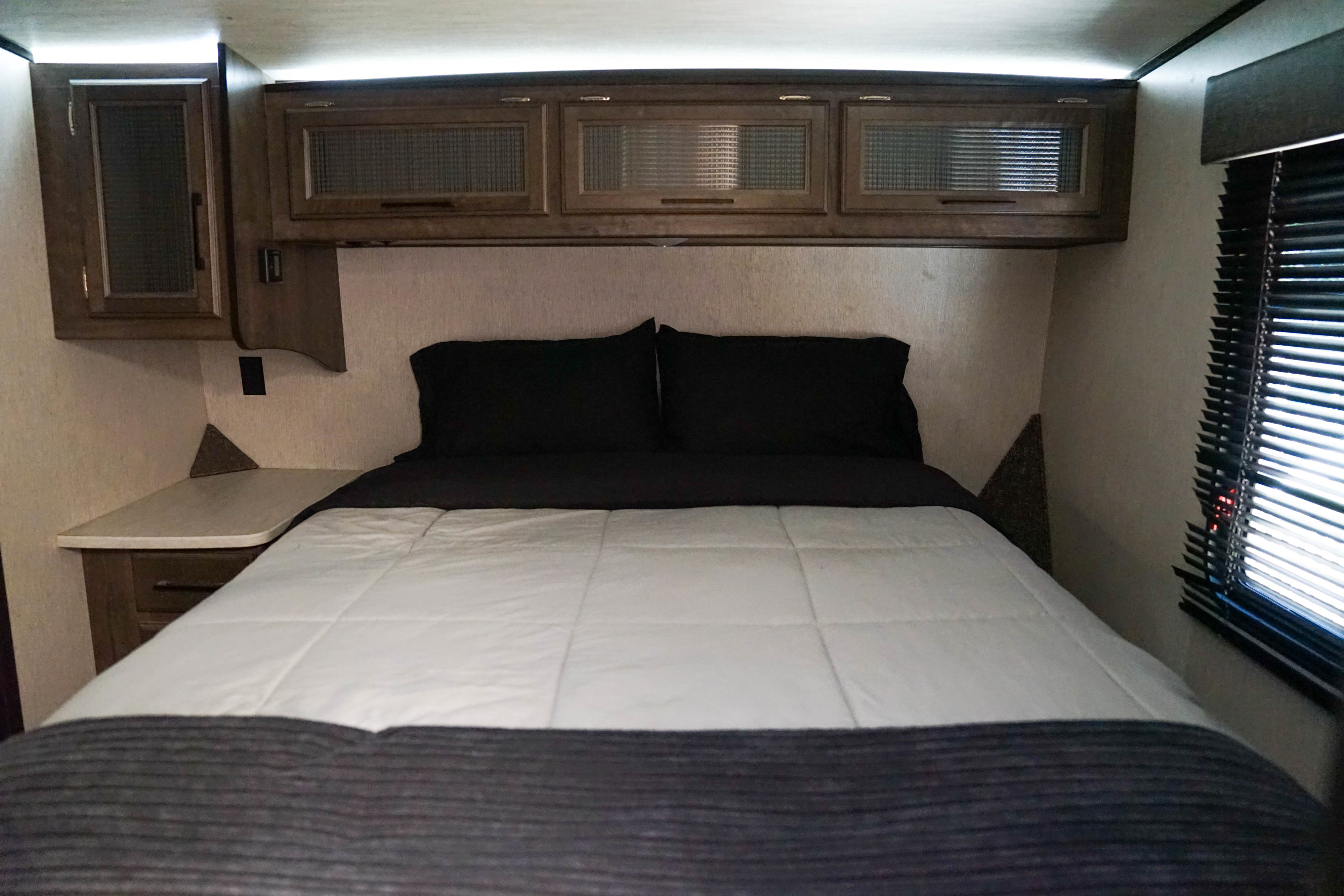 Cozy RV rental bedroom featuring a queen bed, black pillows, and wooden overhead cabinets with ribbed glass.. Heartland RVs Torque Toy Hauler 2019
