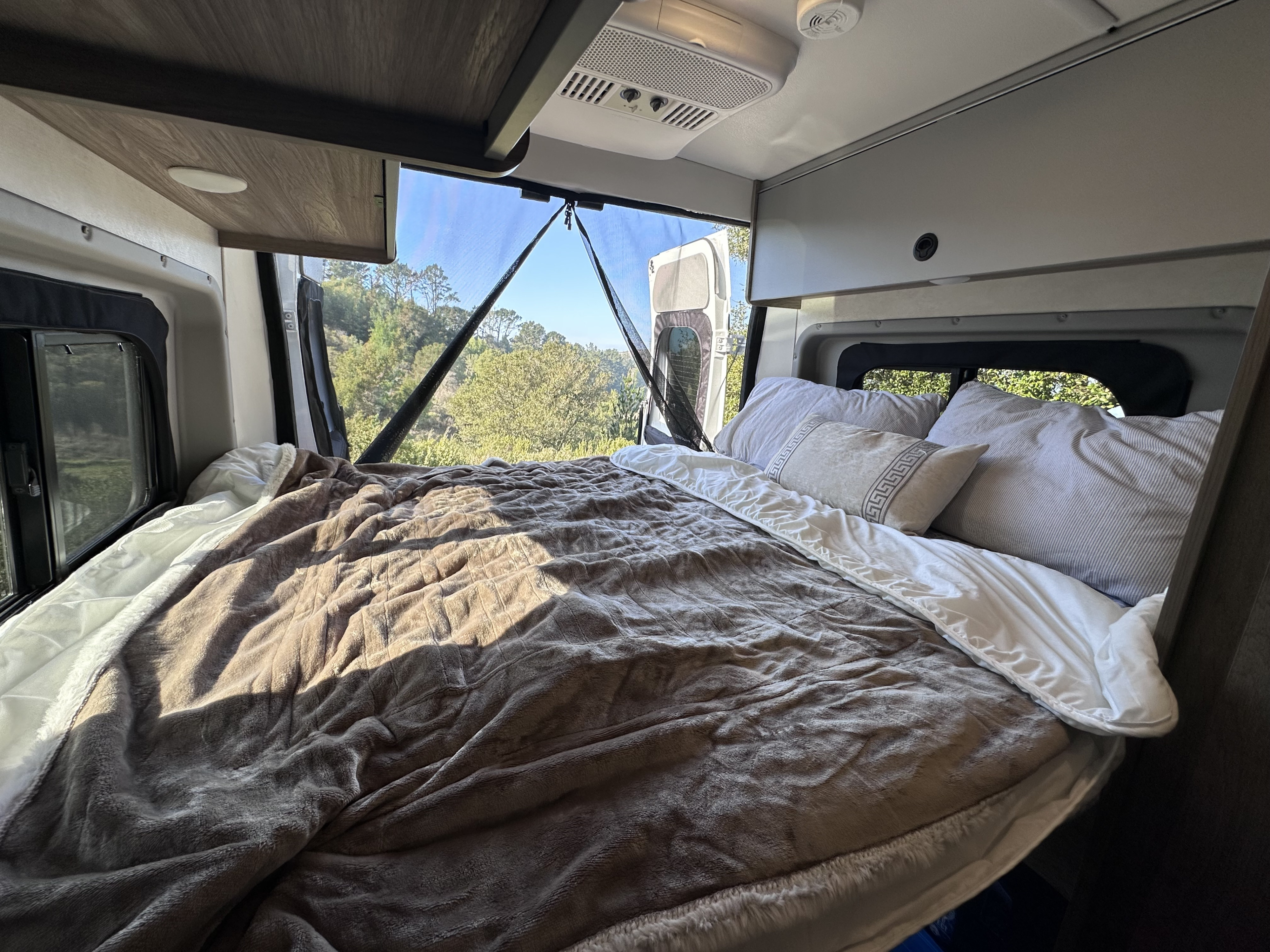 RV rental interior: A cozy bed with a brown blanket and pillows offers serene views of green trees through the back windows.. Winnebago Solis 59P 2026