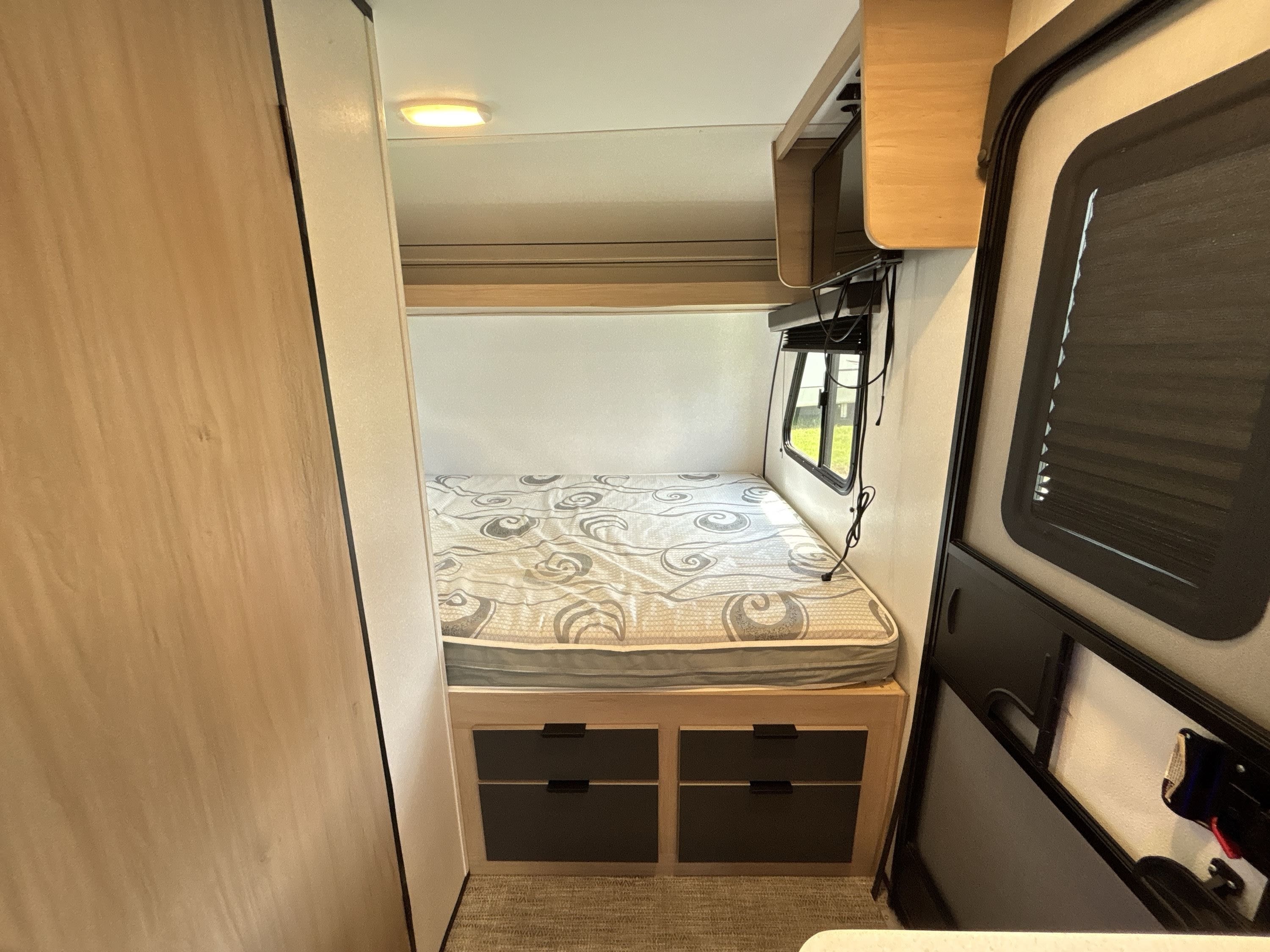 RV rental bedroom with a cozy, patterned bed, under-bed storage, a window, and overhead cabinets.. Cruiser RV Hitch 2021
