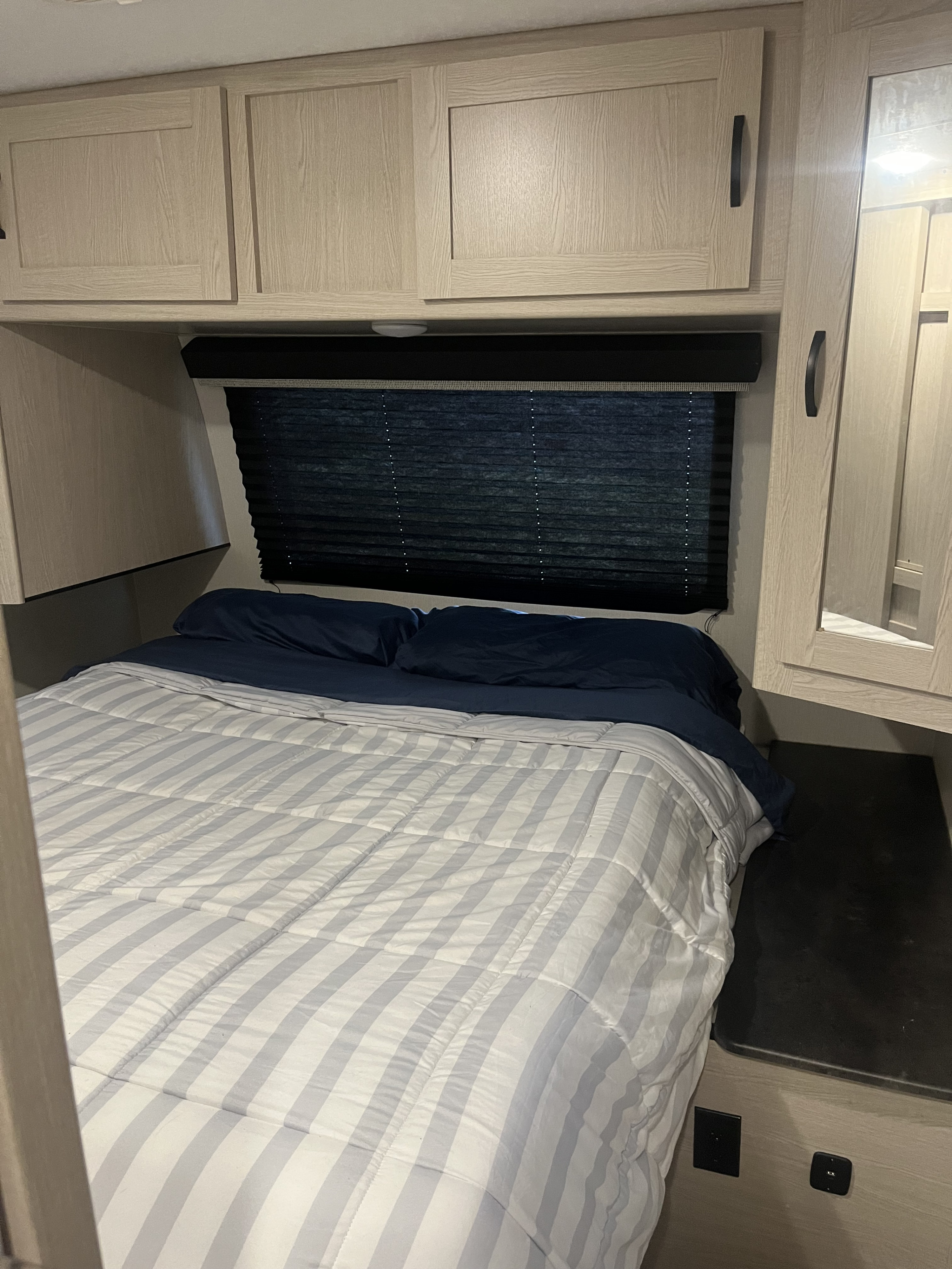 This RV rental bedroom features light wood cabinets, a dark window blind, and a bed with blue sheets and a striped comforter.. Coachmen Apex 2023
