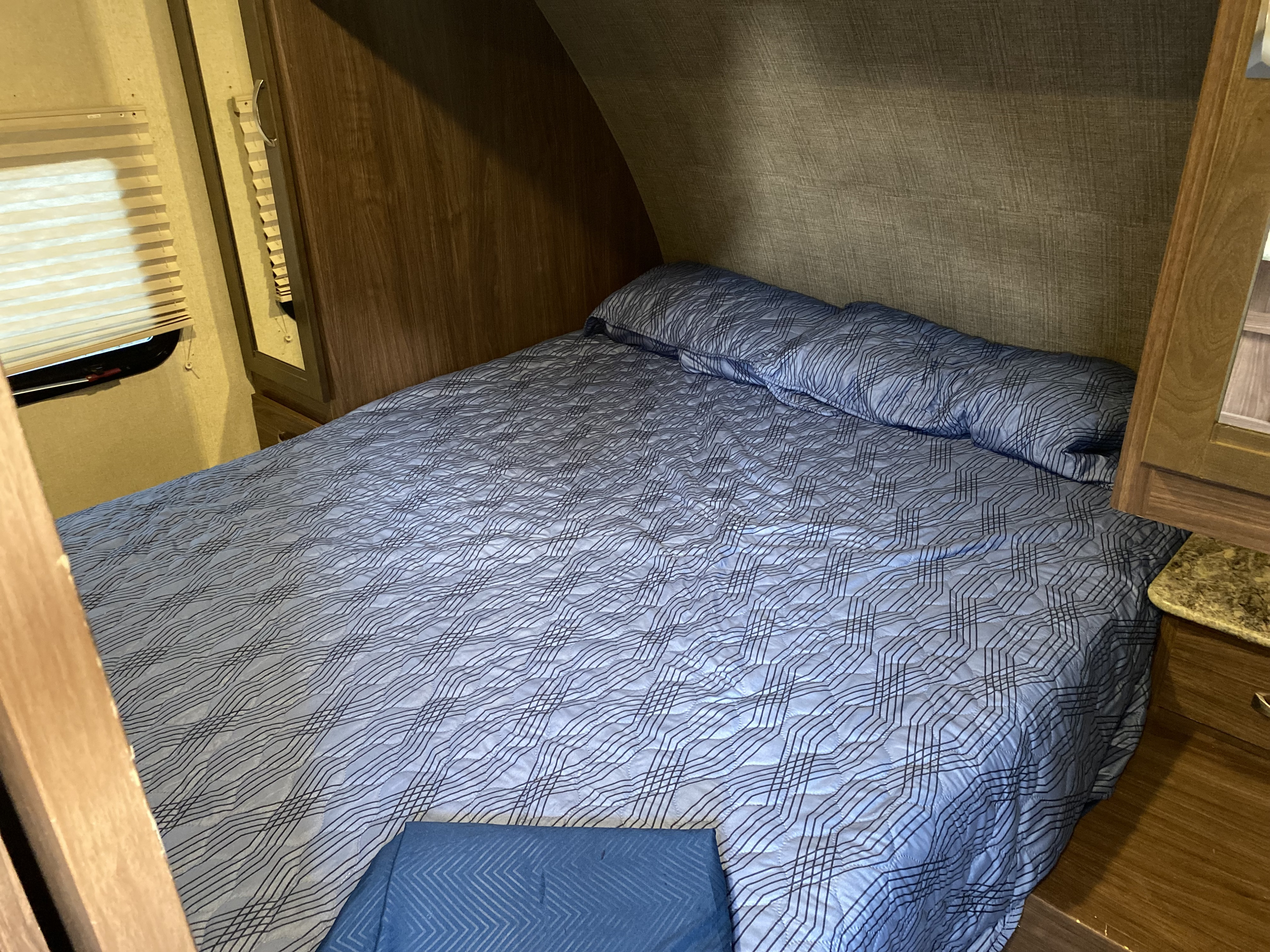 Cozy RV rental bedroom featuring a blue patterned comforter on the bed, window with blinds, and wood paneling.. Coleman 2305QB 2017