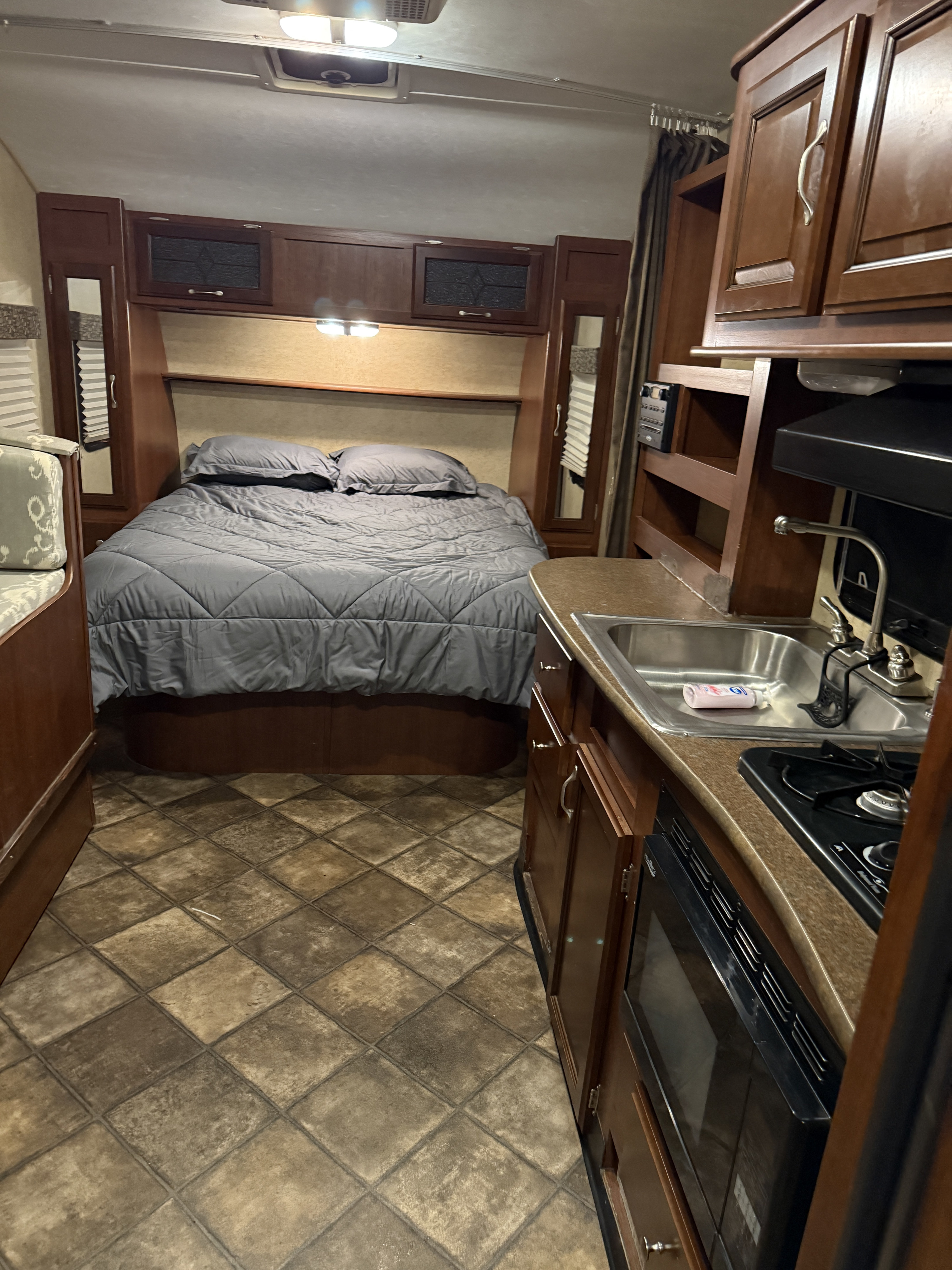 This RV rental interior features a cozy bedroom with gray bedding, a kitchen with a sink & stove, and tiled floors.. Evergreen RV Ascend 2015