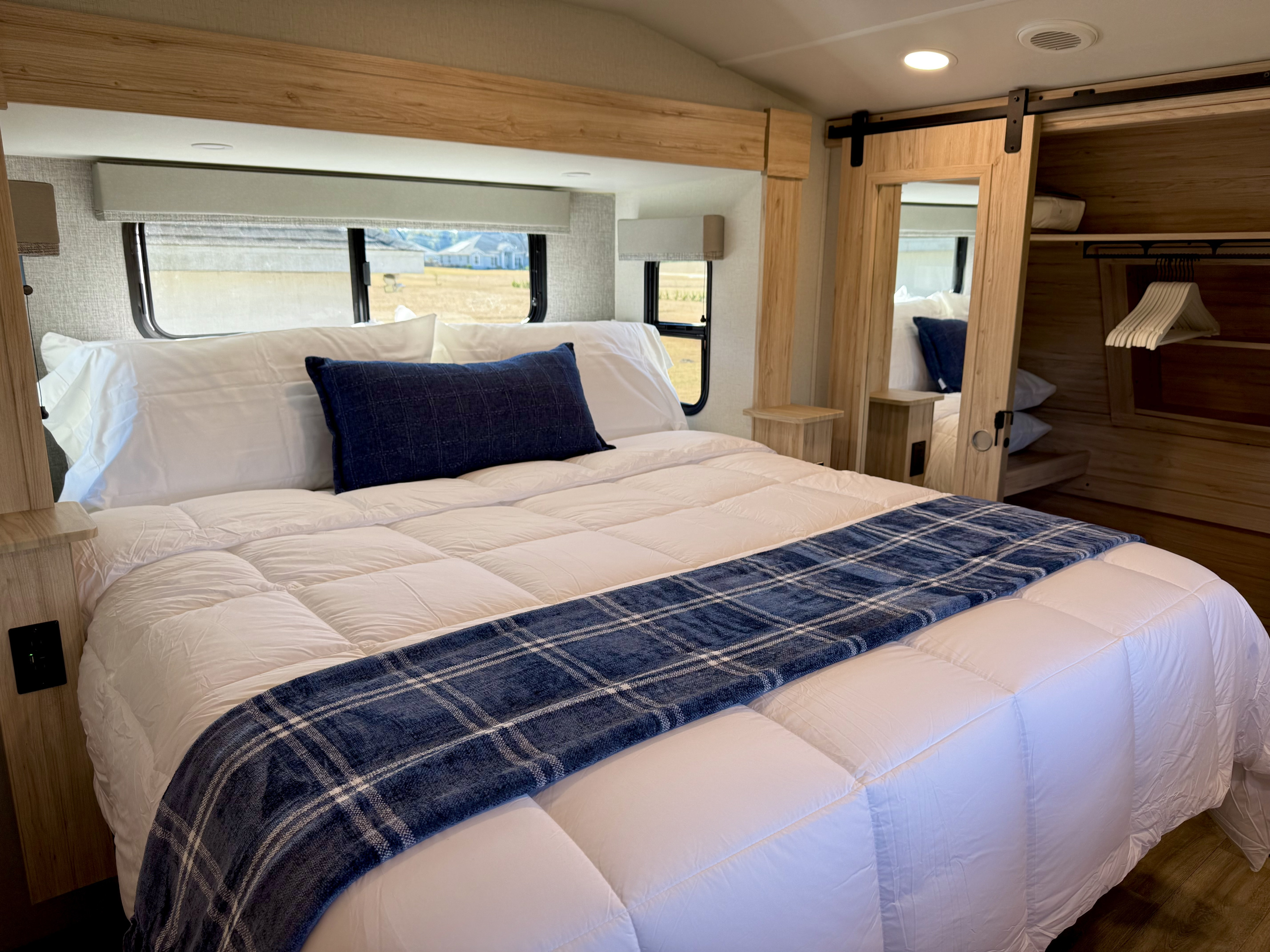 Cozy RV rental bedroom featuring white bedding, blue plaid accents, wooden trim, and views of the outdoors.. Alliance RV Avenue 2023