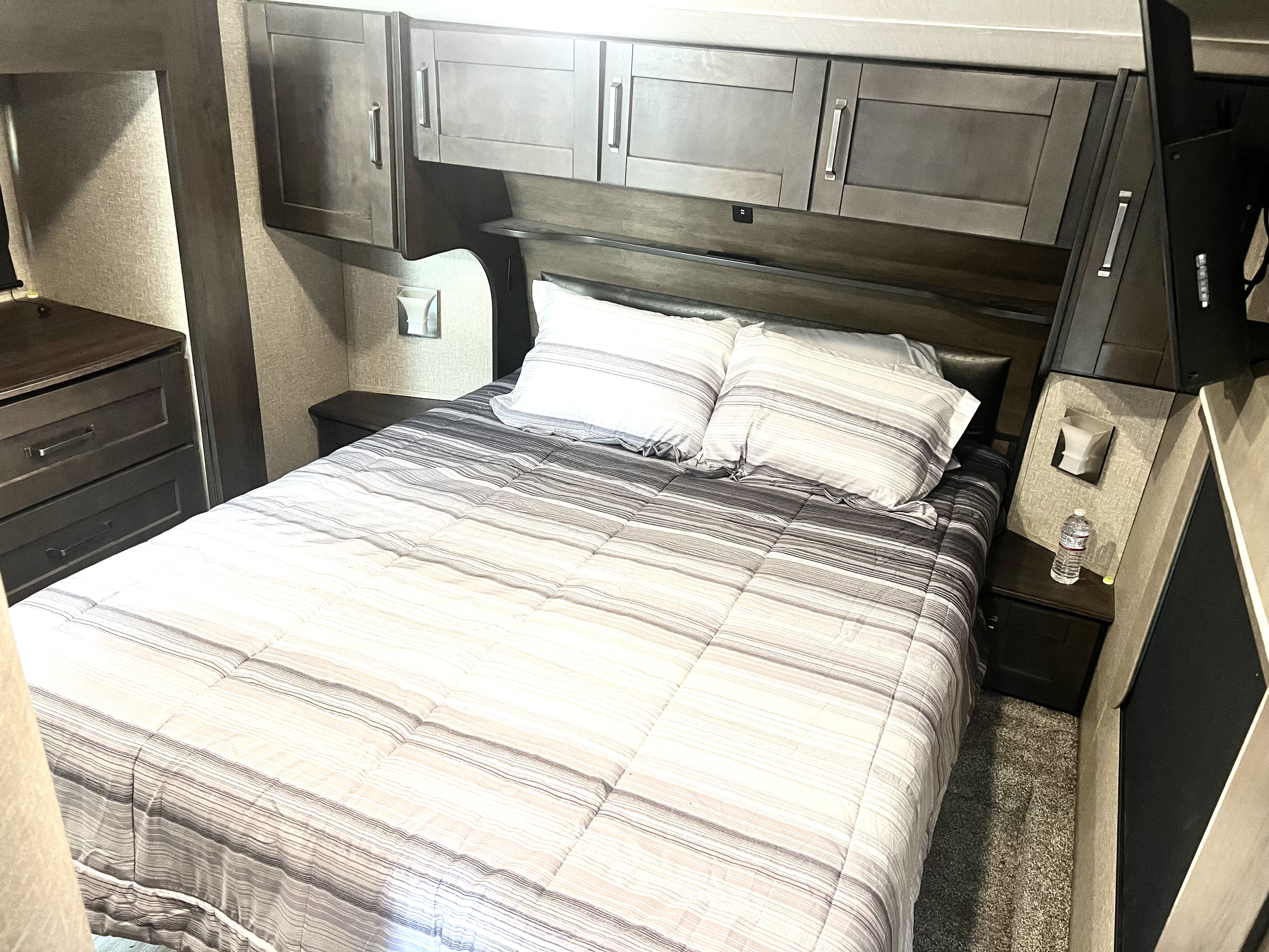 Cozy RV rental bedroom with striped bedding, overhead cabinets, and a nightstand, offering comfortable travel accommodations.. Grand Design Reflection 2021
