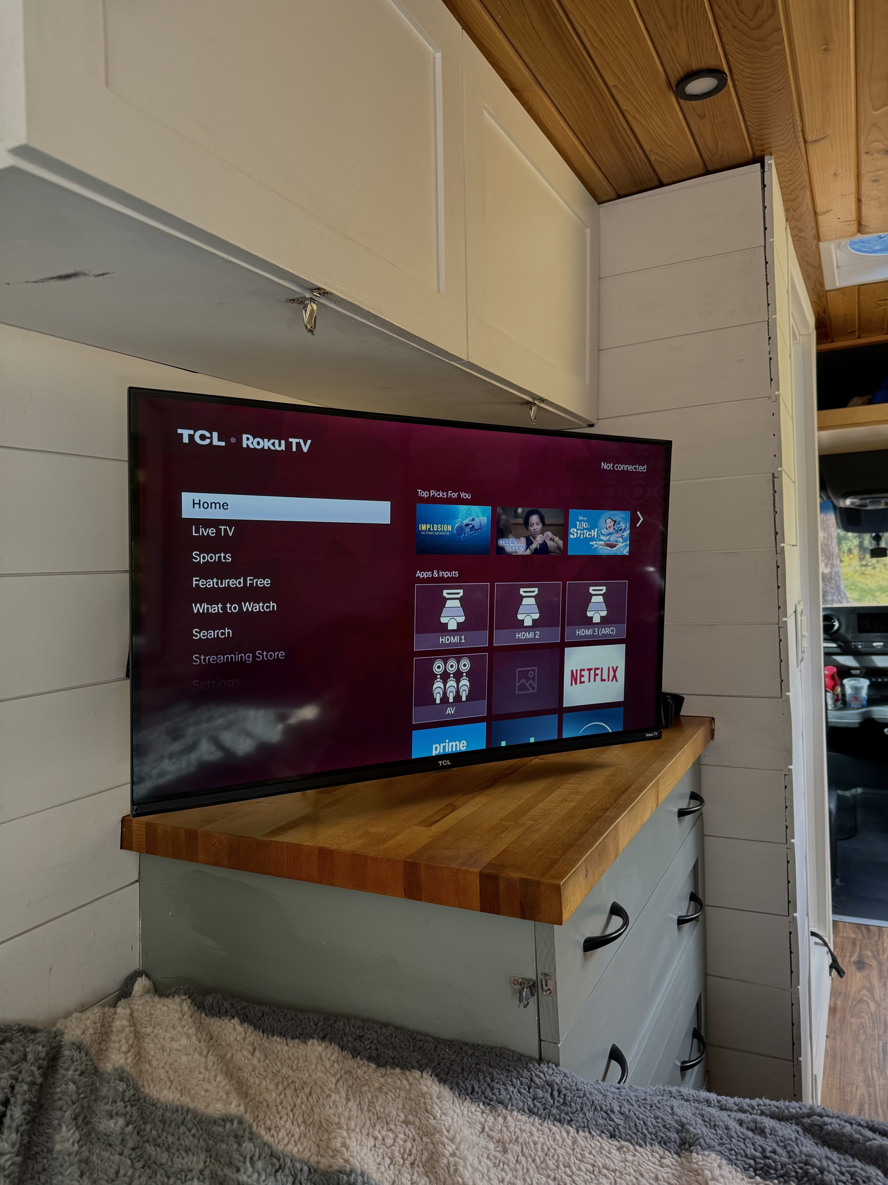 This RV rental features a TCL Roku TV displaying streaming apps and inputs on a wooden counter, part of a cozy custom-built interior.. Mercedes-Benz Sprinter 2020
