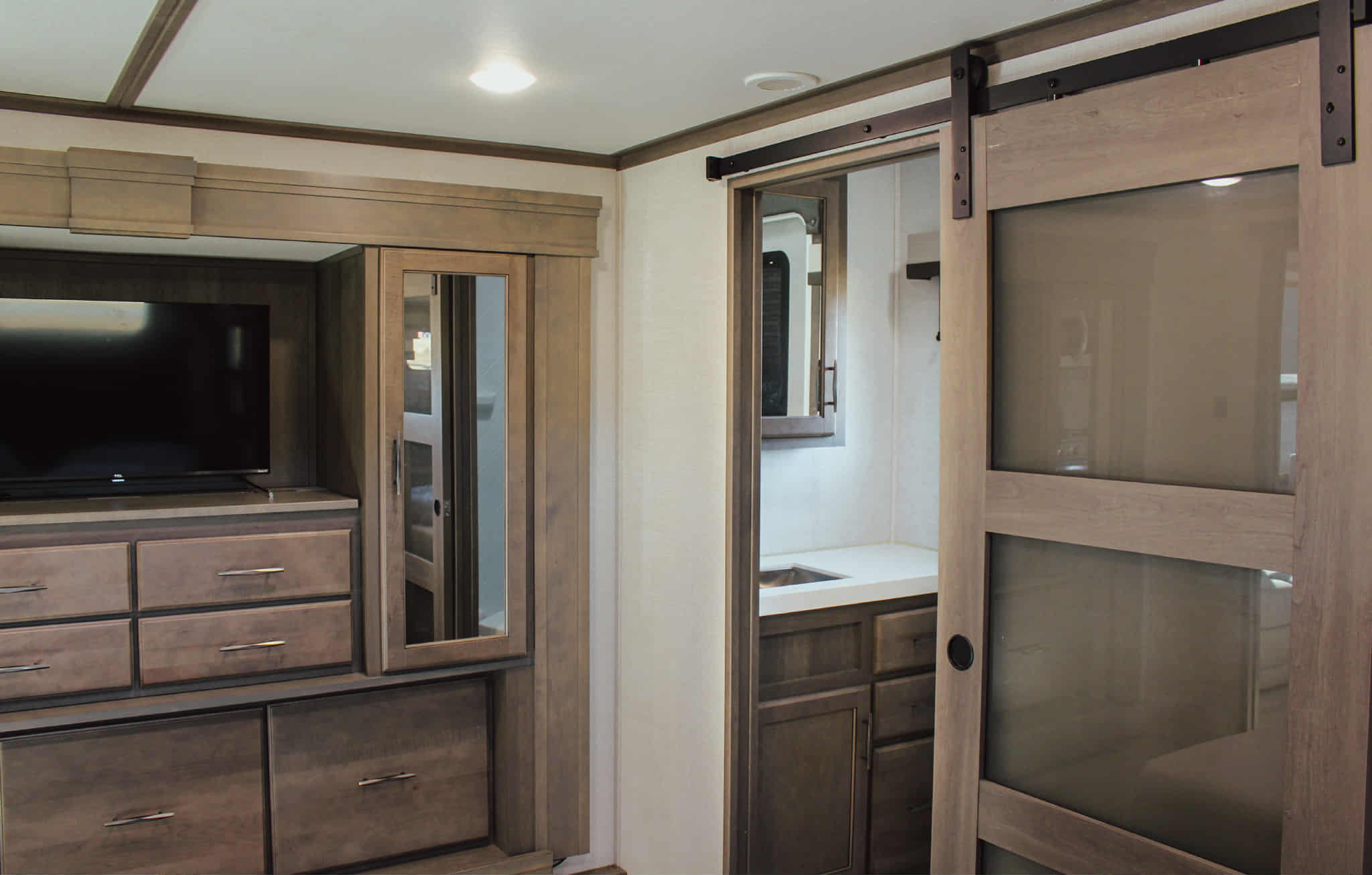 Modern rv rental interior with a bedroom TV, storage drawers, and a sliding barn door leading to a well-equipped bathroom.. Grand Design Solitude 2023