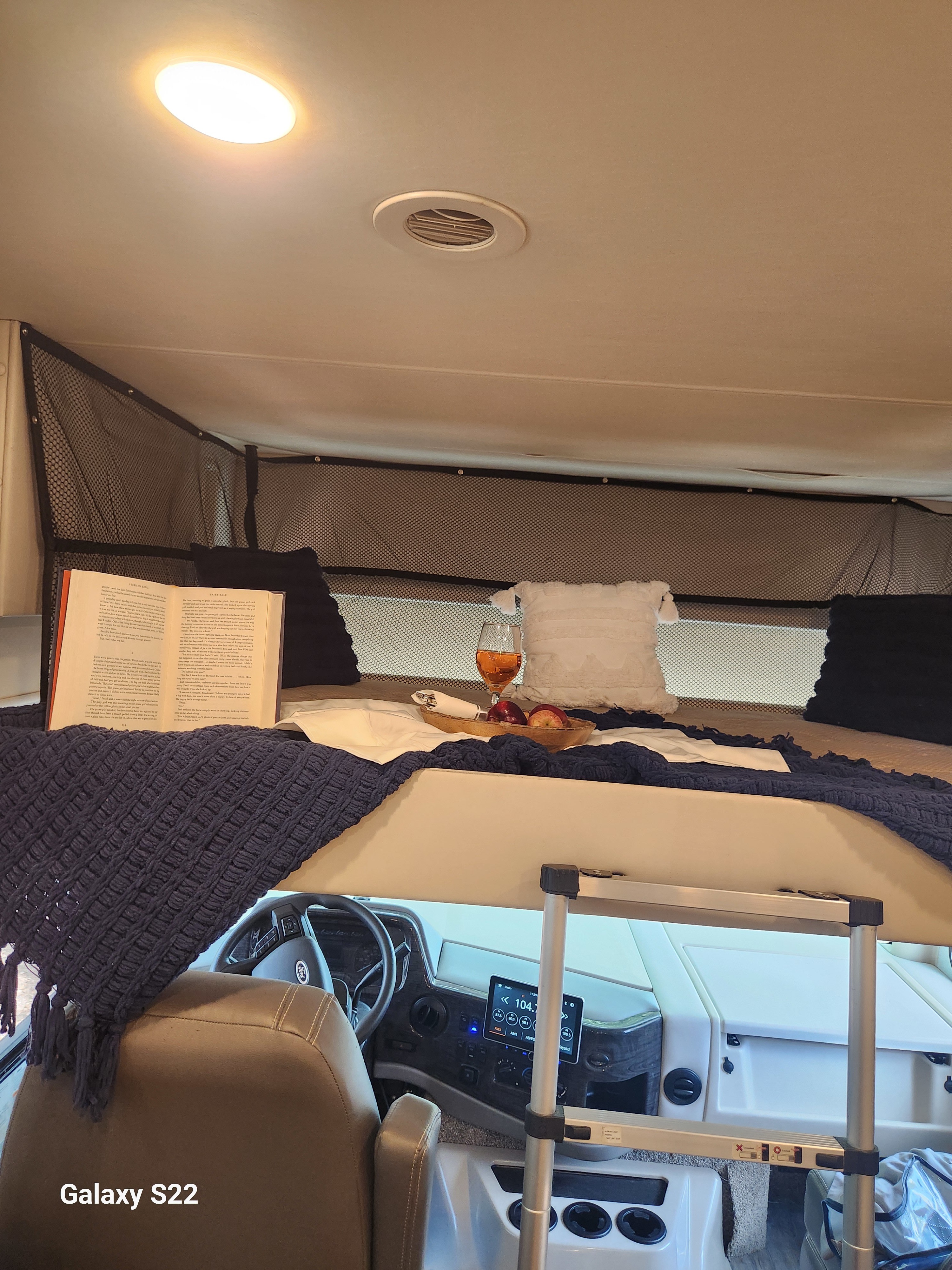 This RV rental features a cozy over-cab bunk bed with a book, drink, and fruit, visible above the driver's cabin.. Thor Miramar 2023