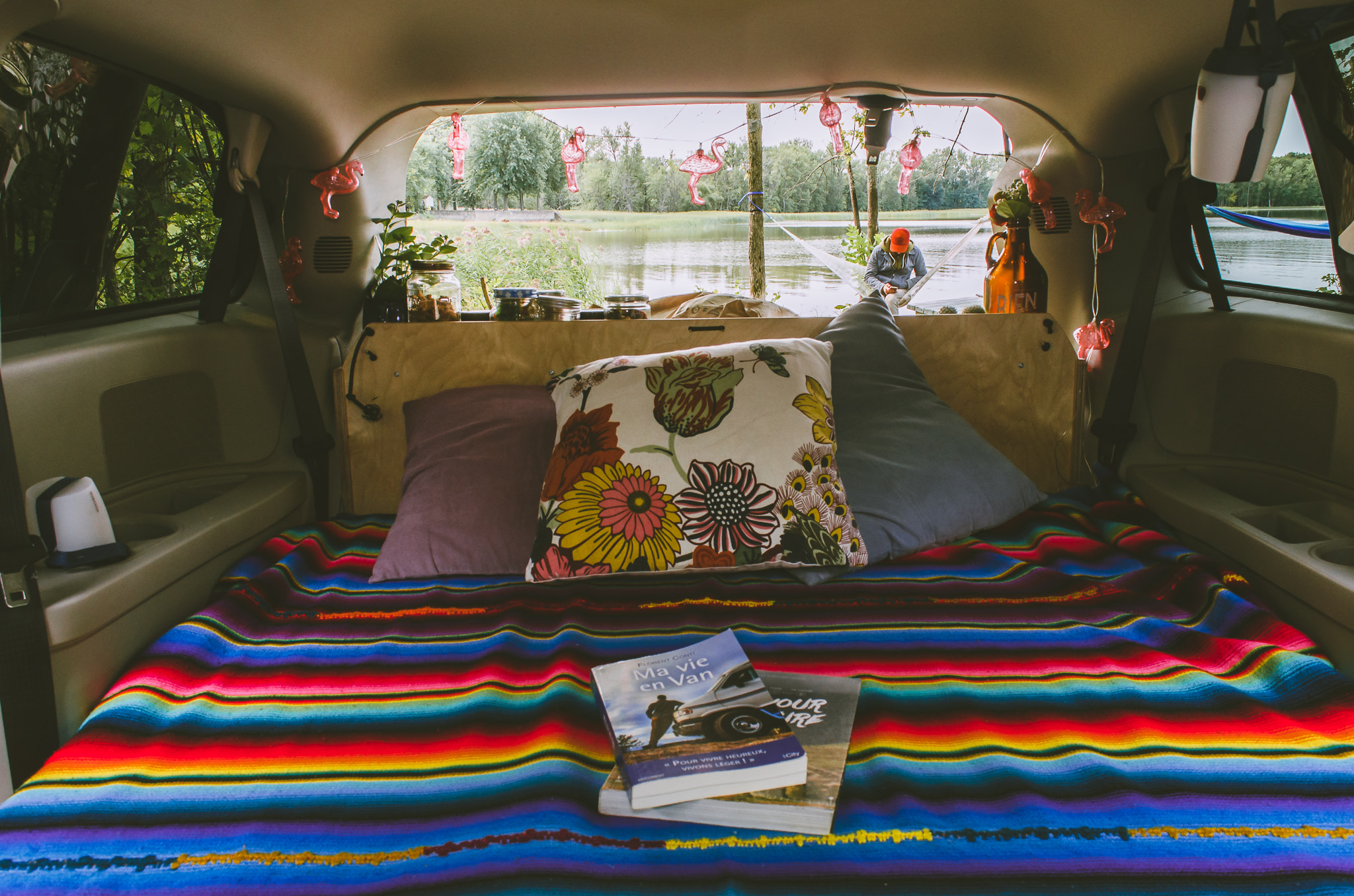 Relax in a cozy RV rental van with a vibrant bed, books, and a beautiful lake view from the open back.. chrysler Grand Caravan 2024