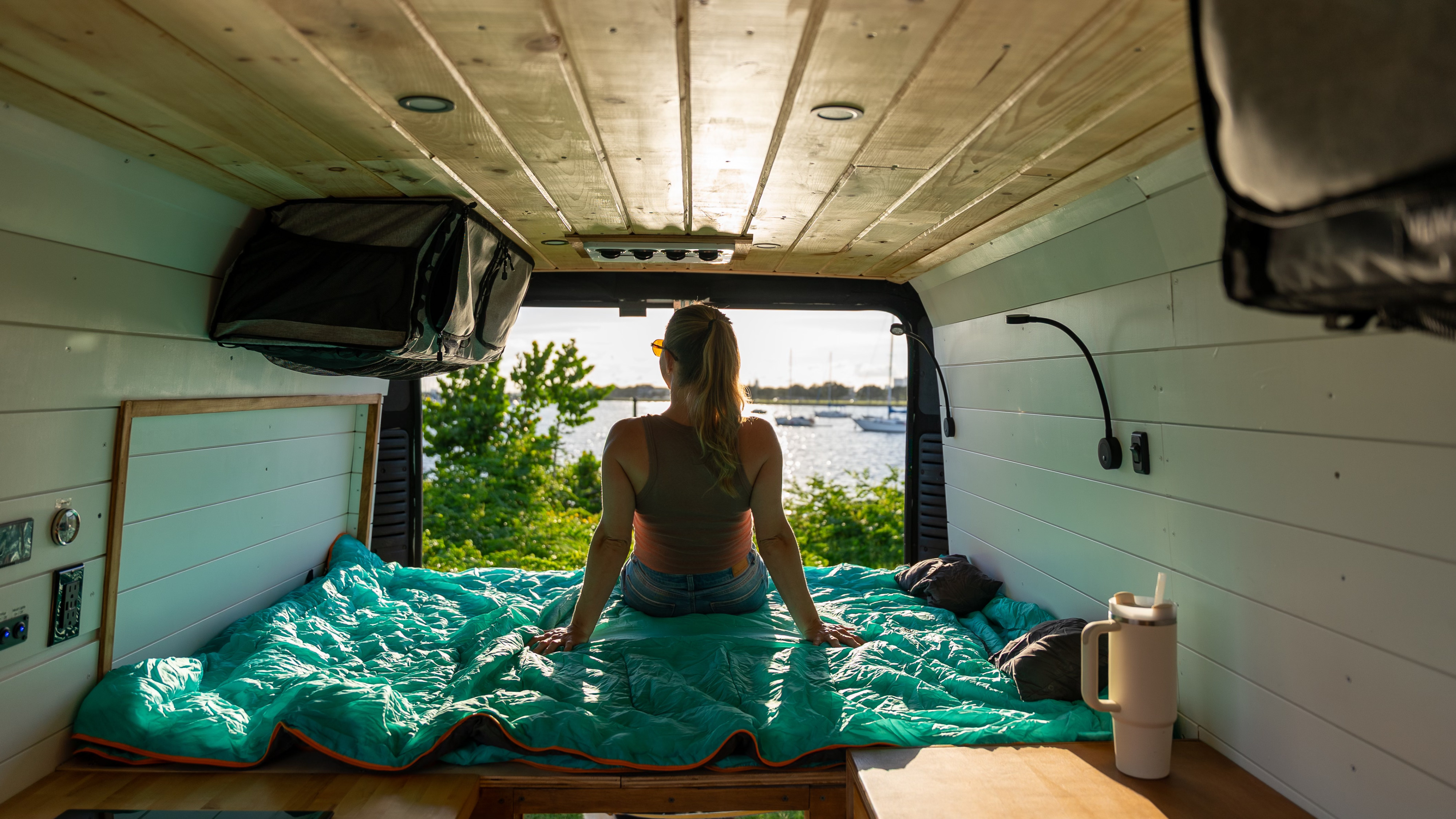 A woman sits on a turquoise sleeping bag in a cozy RV rental van, looking out at boats on the water.. Ram Promaster 2500 2021
